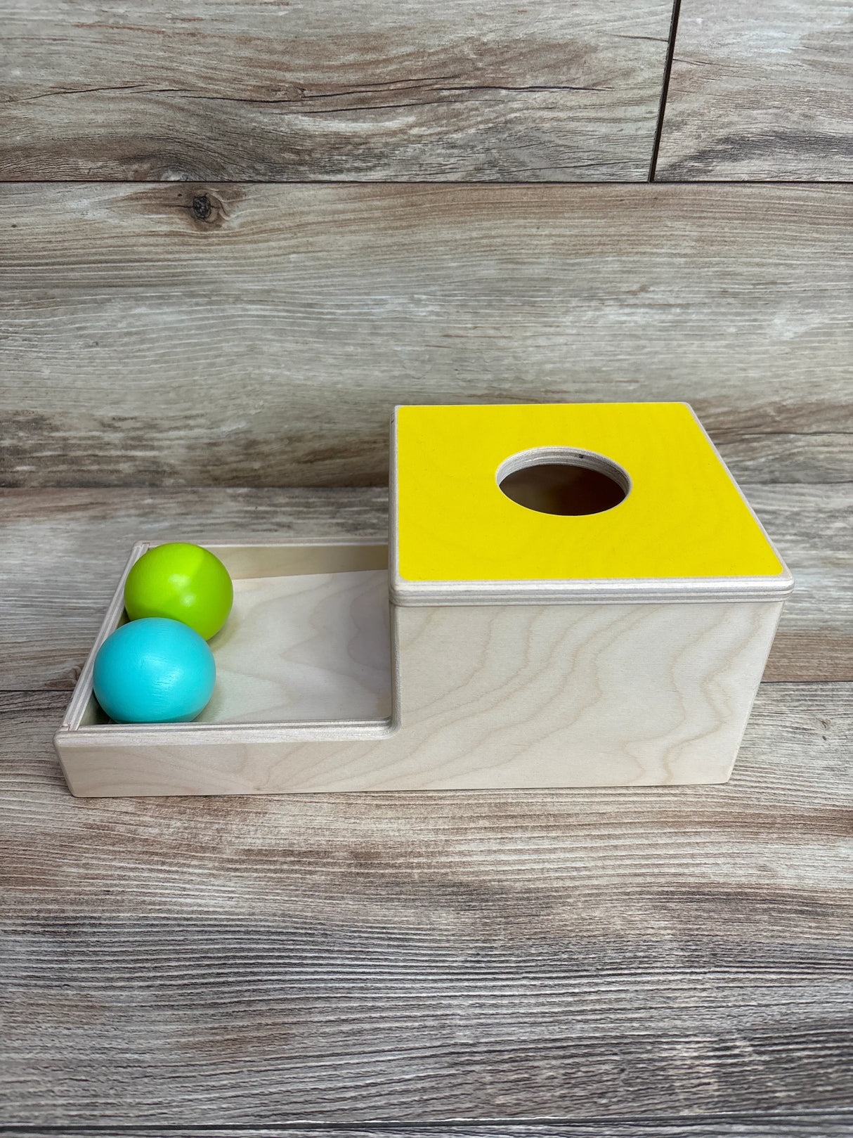 Wooden toddler toy with yellow top, hole for balls, and two colorful balls on wooden surface