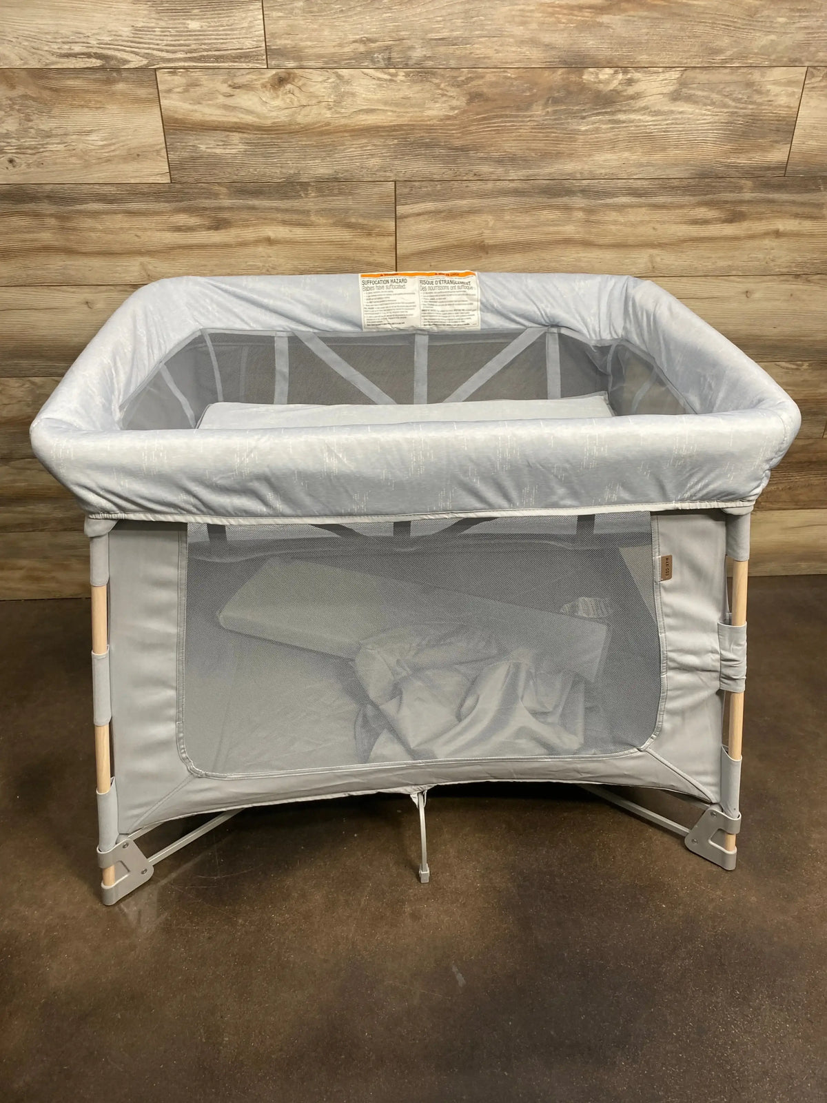 Front view of Maxi-Cosi Swift playard showing gray fabric, mesh sides, and wooden-look legs on a wood-paneled floor