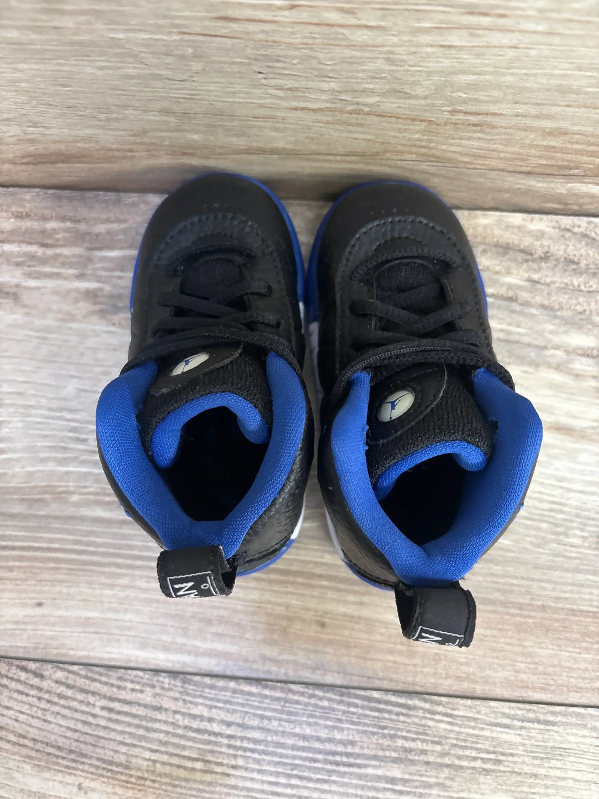 Top view of Air Jordan Jumpman Pro BT toddler sneakers showing black pebbled uppers, blue padded collars and Jumpman logos on the tongues on a light wood background