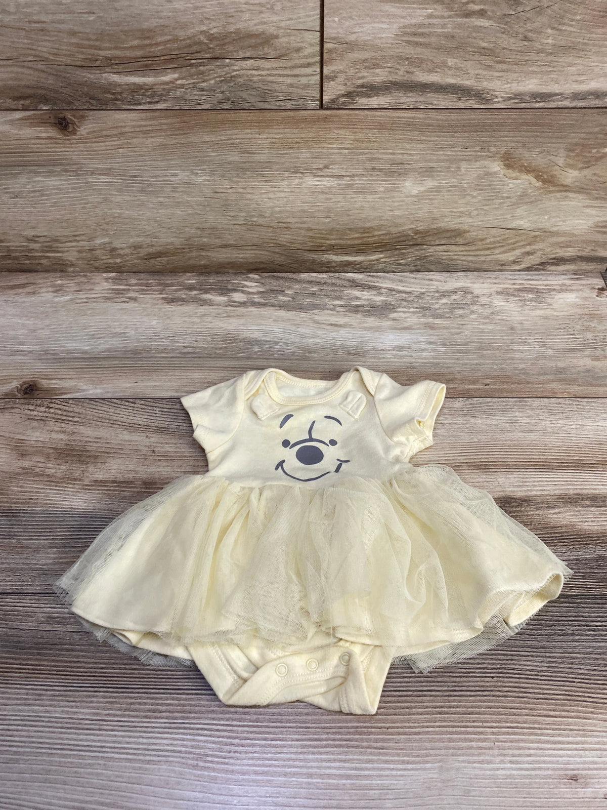 Front view of Disney Baby Winnie the Pooh yellow tutu bodysuit showcasing Pooh face graphic and layered tulle skirt on a wood surface