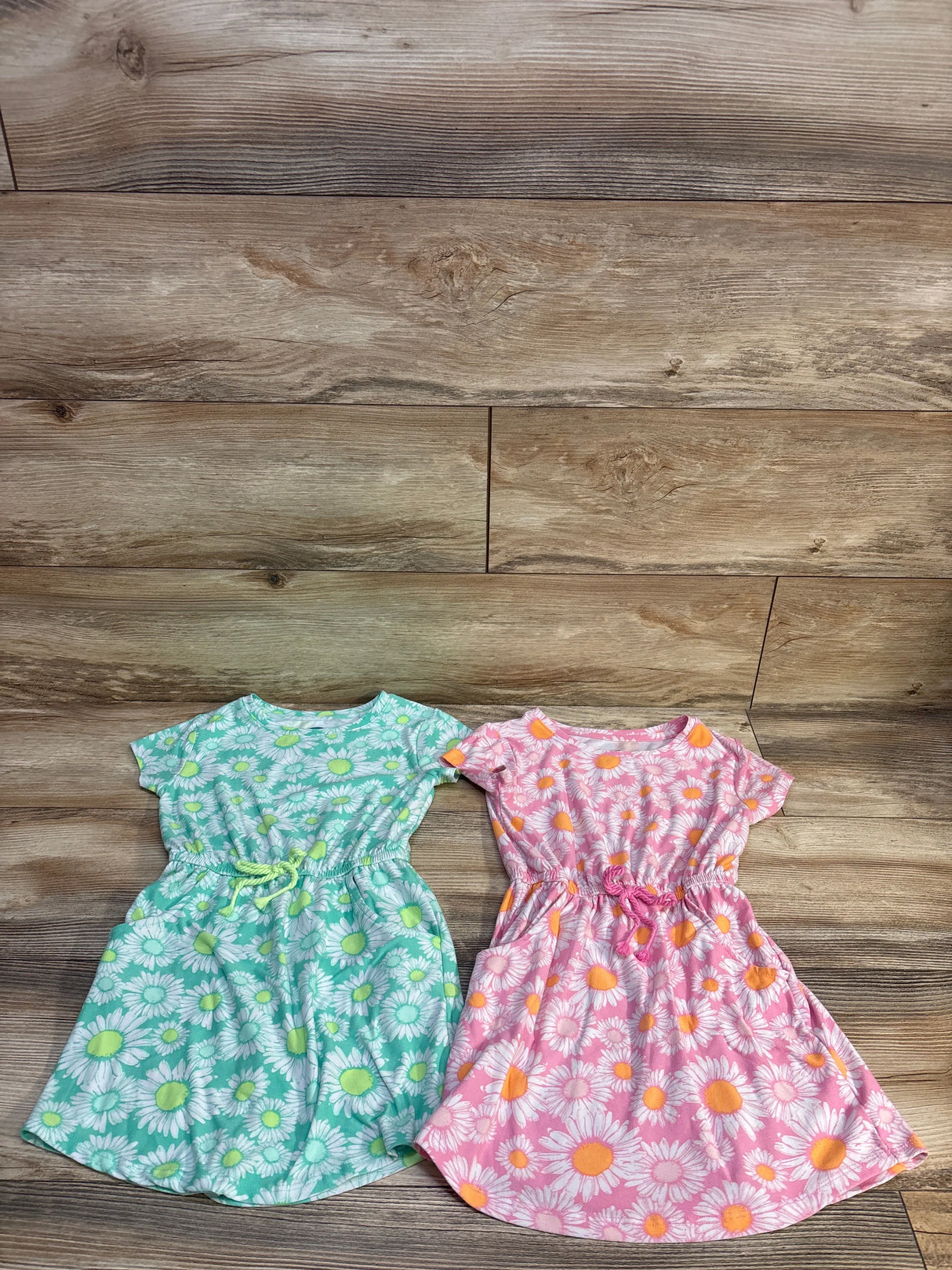 Flat lay of two toddler floral dresses, green and pink, showing daisy print and elastic cinched waists on wood floor