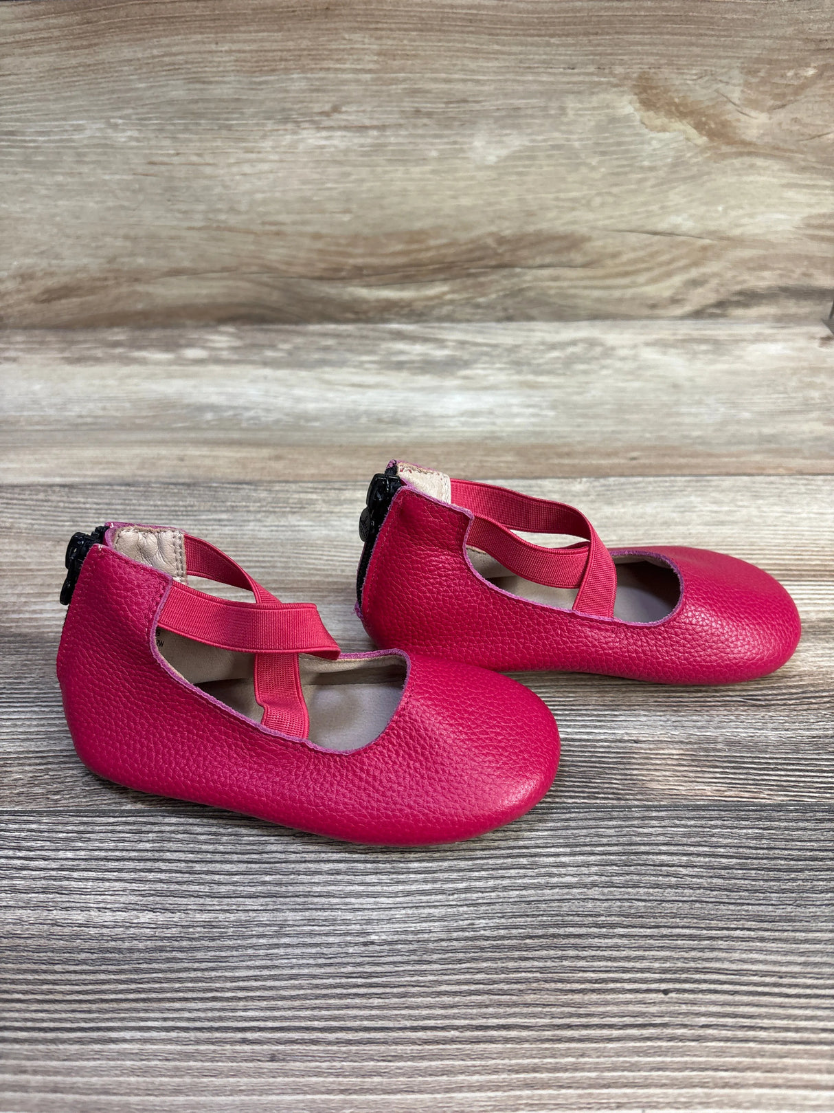 Side view of pink Mary Jane baby ballet flats showing criss-cross elastic straps and back zipper on wood tabletop