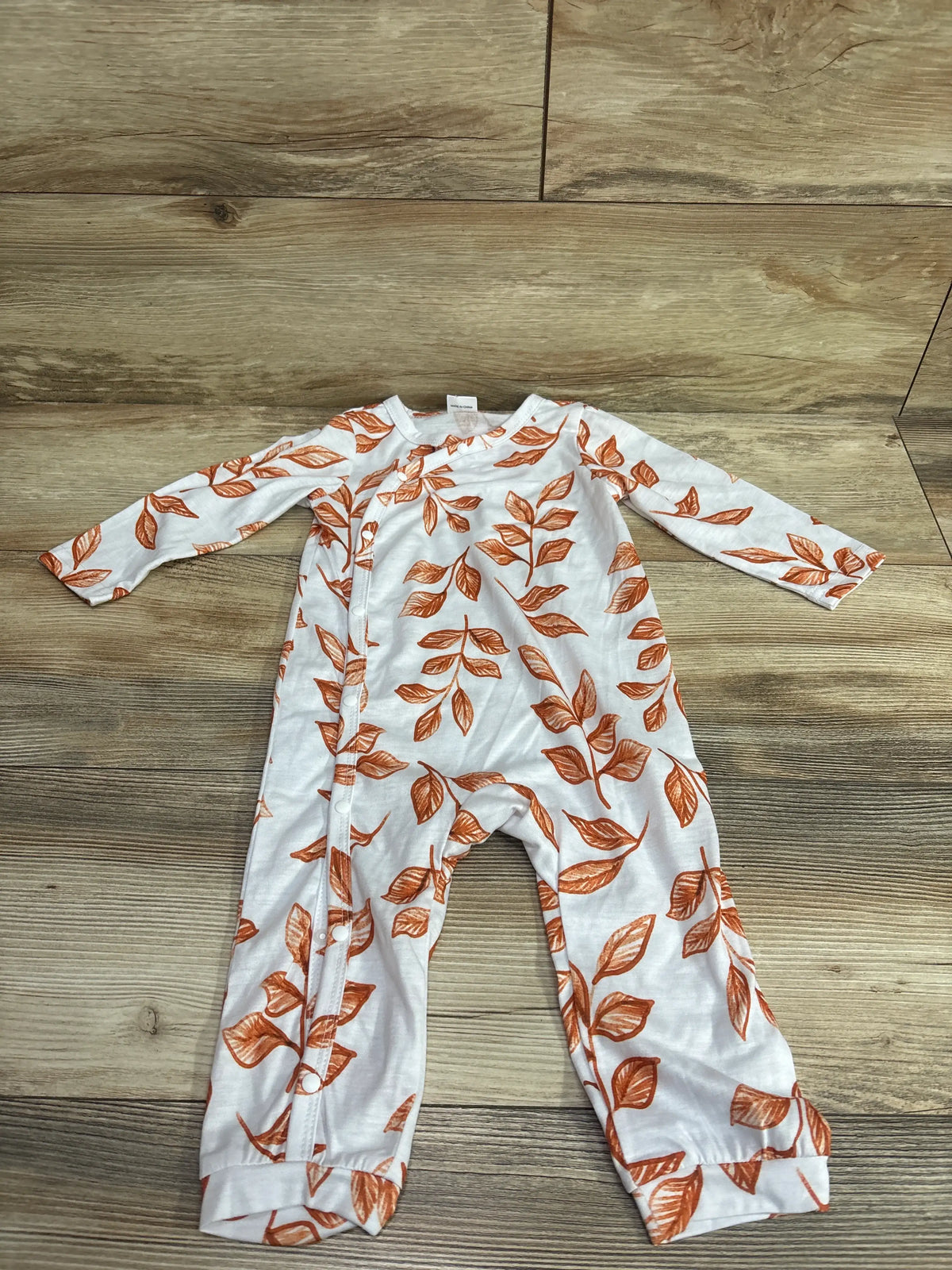 Front view of a white PatPat baby coverall showcasing a brown leaf print and side snap closure on a wood floor