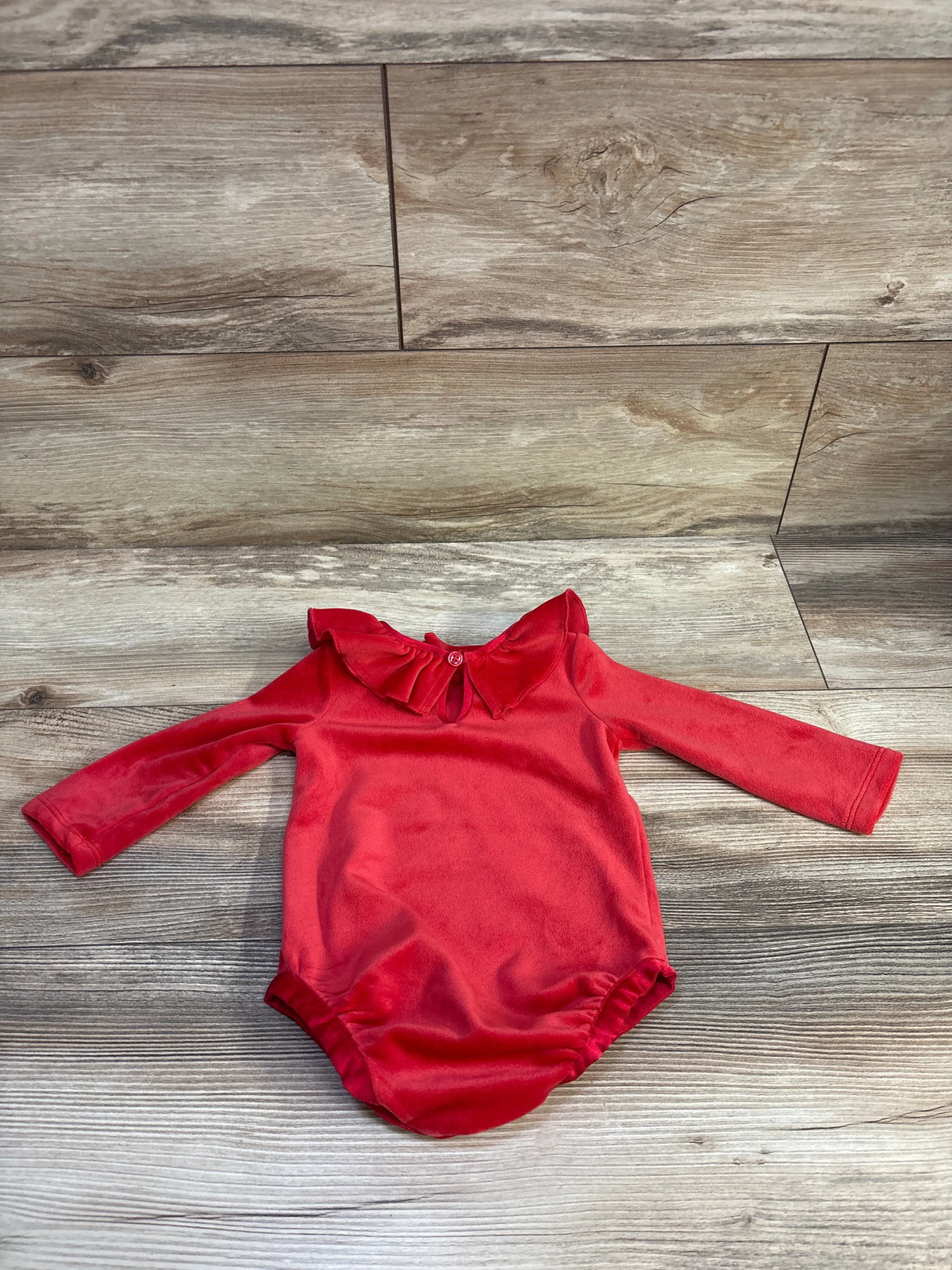 Flat lay of red velour collared long-sleeve baby bodysuit showing ruffled collar and back button on wood background