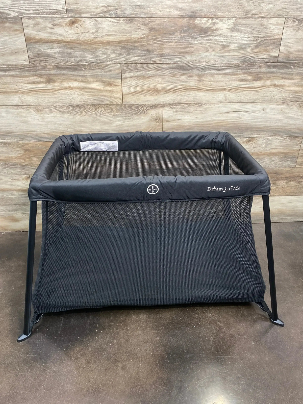Front view of Dream On Me Travel Light playard in black showcasing mesh sides and metal legs against a wood-paneled wall background
