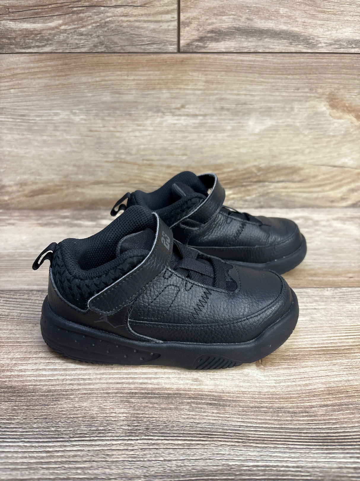 Side view of Jordan Max Aura 3 TD Black Anthracite toddler sneakers showing textured black leather upper, Velcro strap, pull tab and cushioned sole on wooden floor