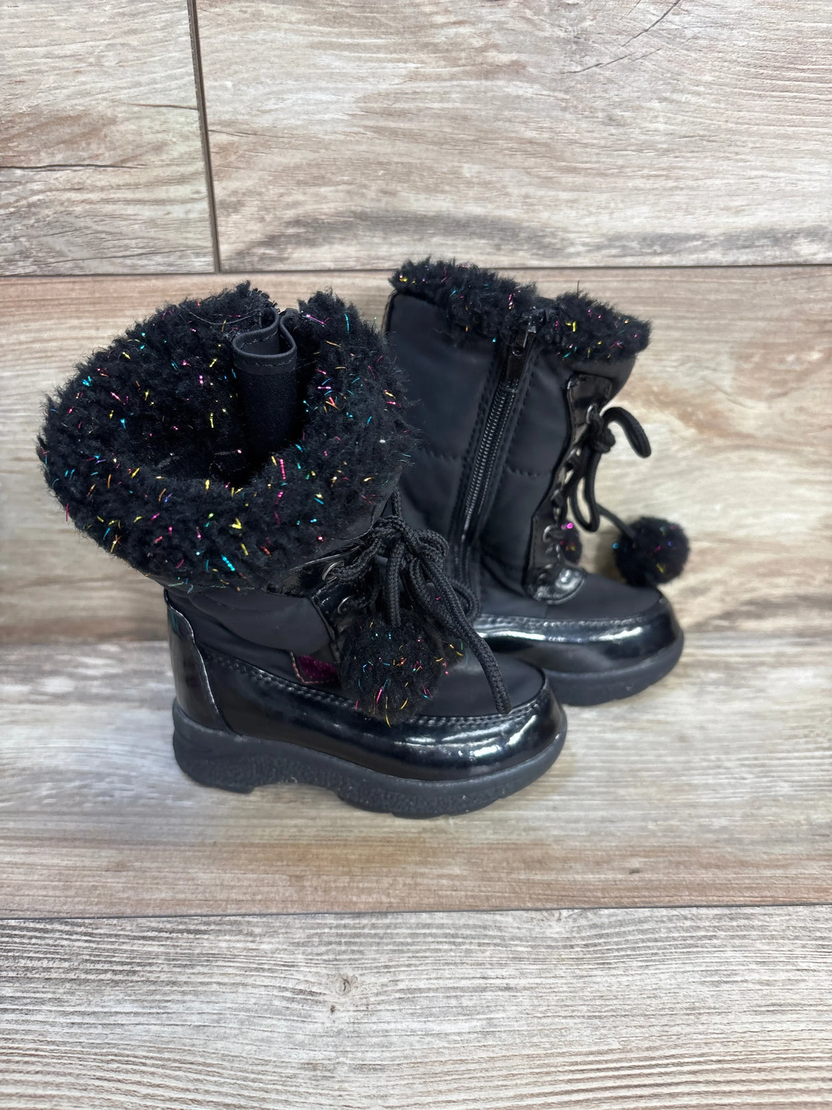 Black toddler winter boots with faux fur trim and pom-pom details on a wooden floor.
