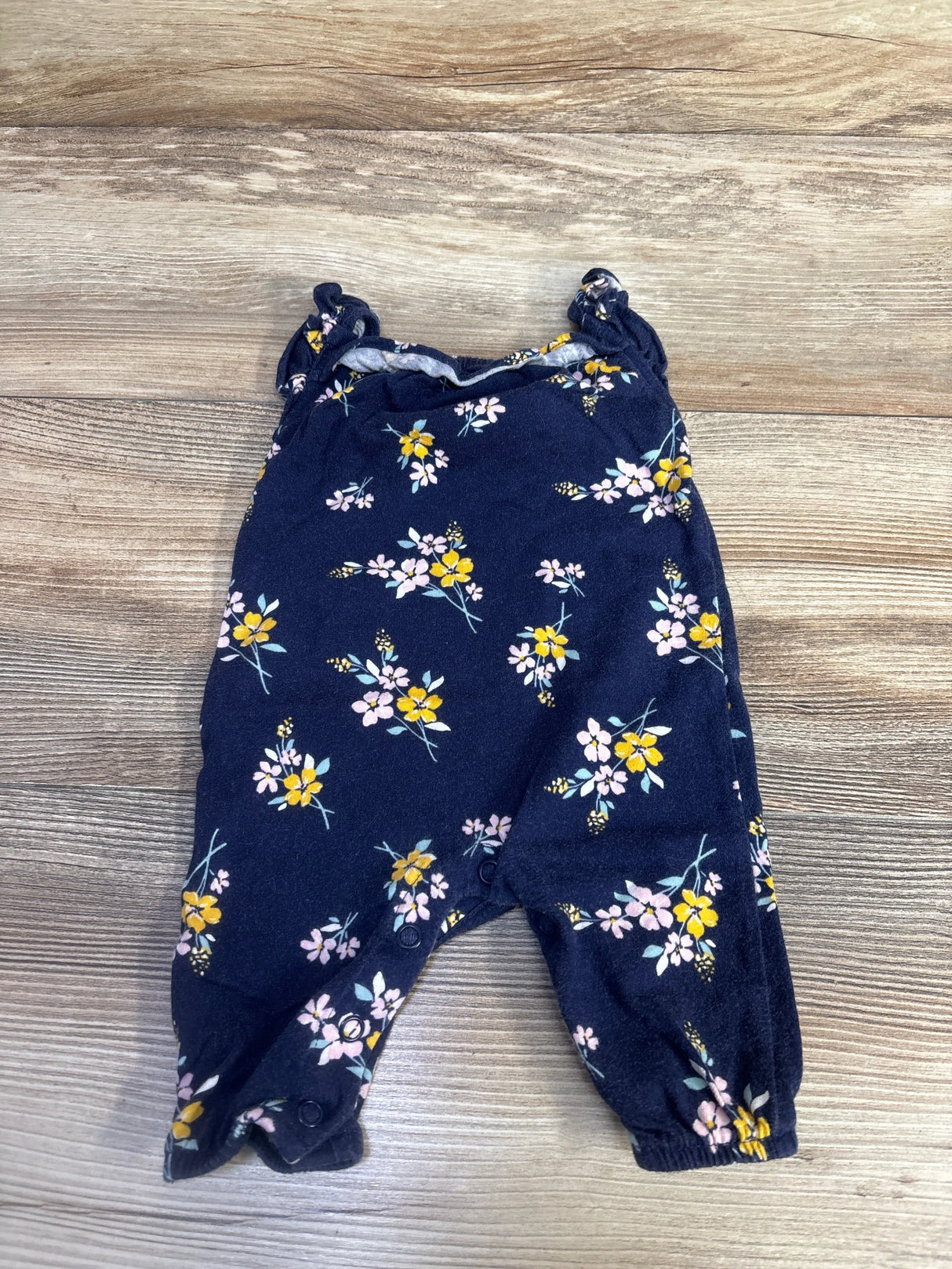 Flat-lay of navy floral sleeveless newborn jumpsuit showing ruffled shoulder straps and snap crotch on wood background