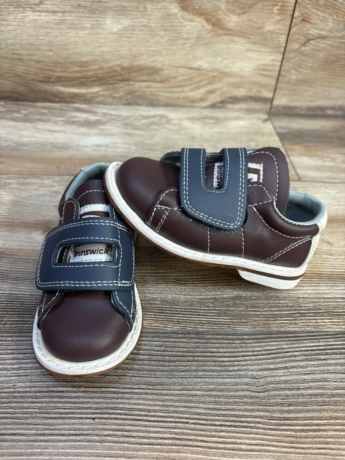 Angled view of Brunswick bowling sneakers showcasing brown uppers, navy hook and loop closures, and white soles on a wood-look floor