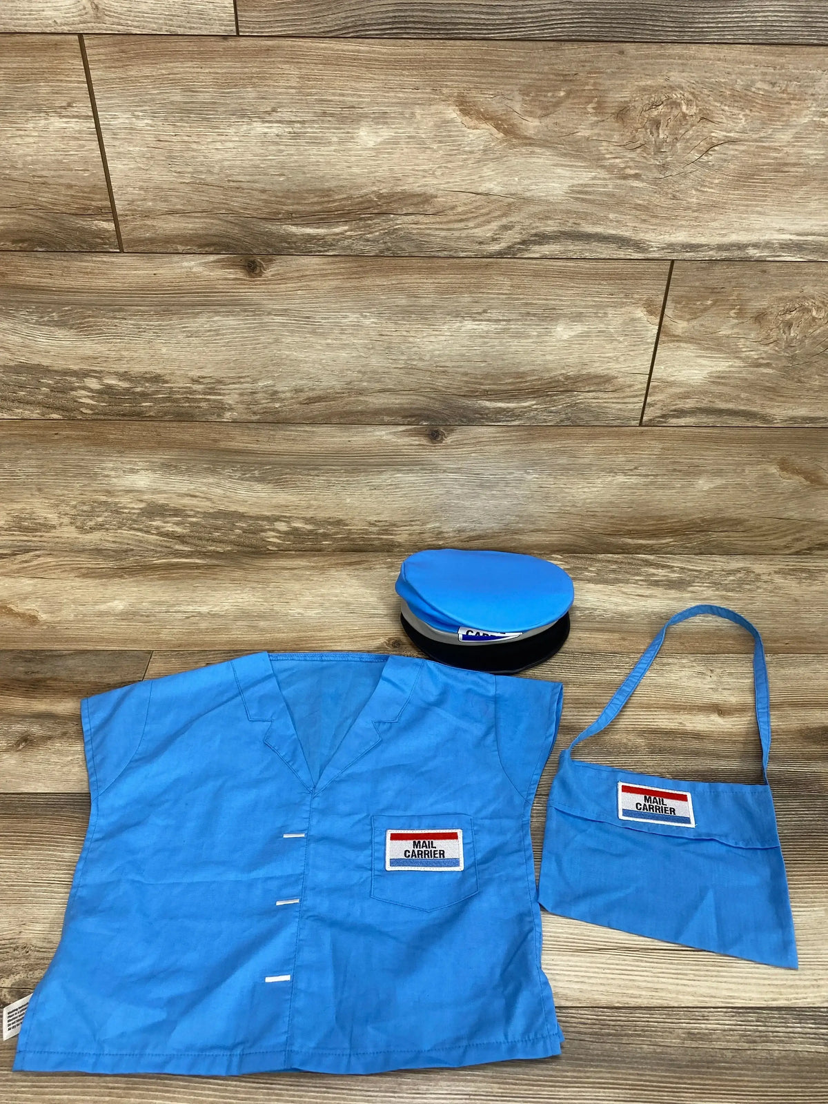 Top-down view of Lakeshore Toddler Mail Carrier Costume showcasing blue shirt, cap, and shoulder bag with Mail Carrier labels on a wood floor