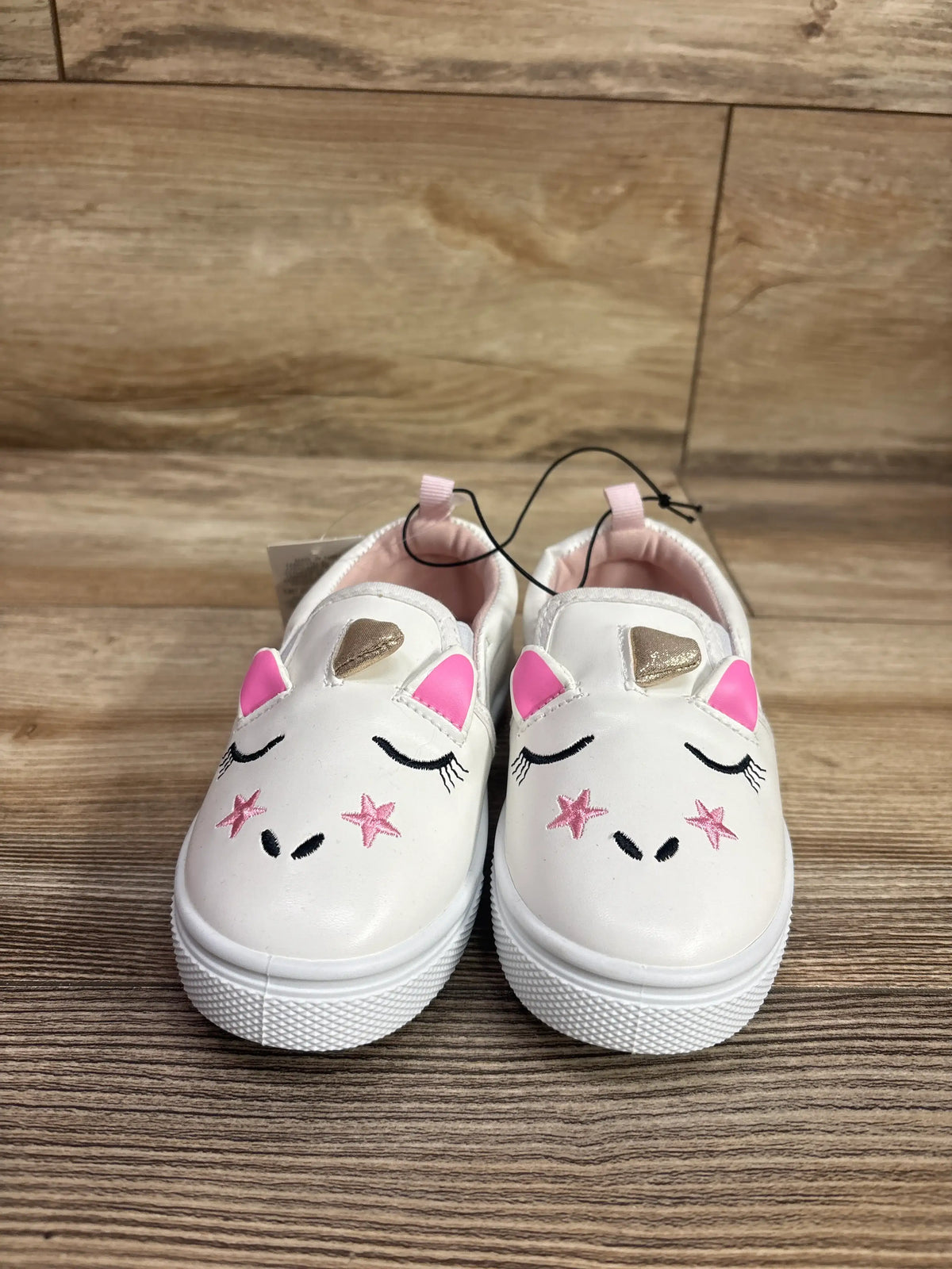Front view of white Bocca slip-on unicorn sneakers showcasing pink ears, gold horns, and embroidered faces on wood flooring