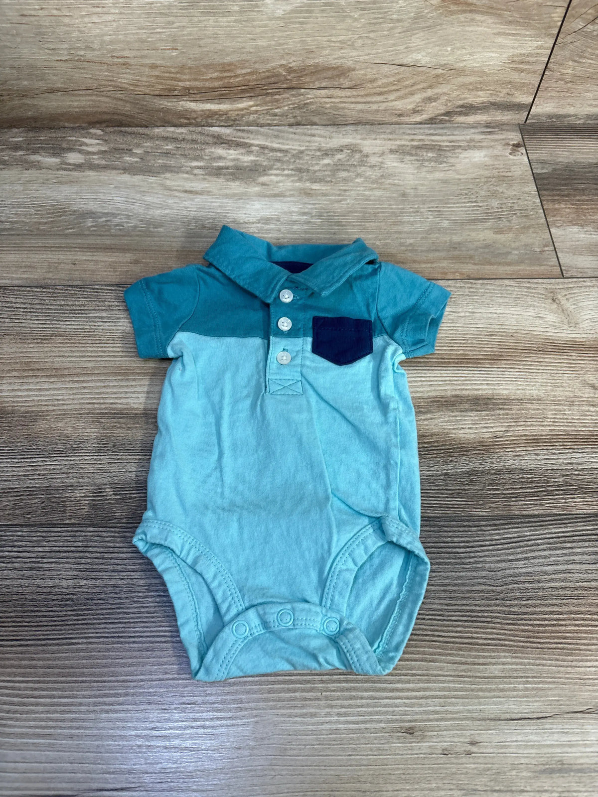 Front view of Carter’s blue newborn polo bodysuit showcasing collar, button closure, chest pocket, and snap closure on wood-look flooring