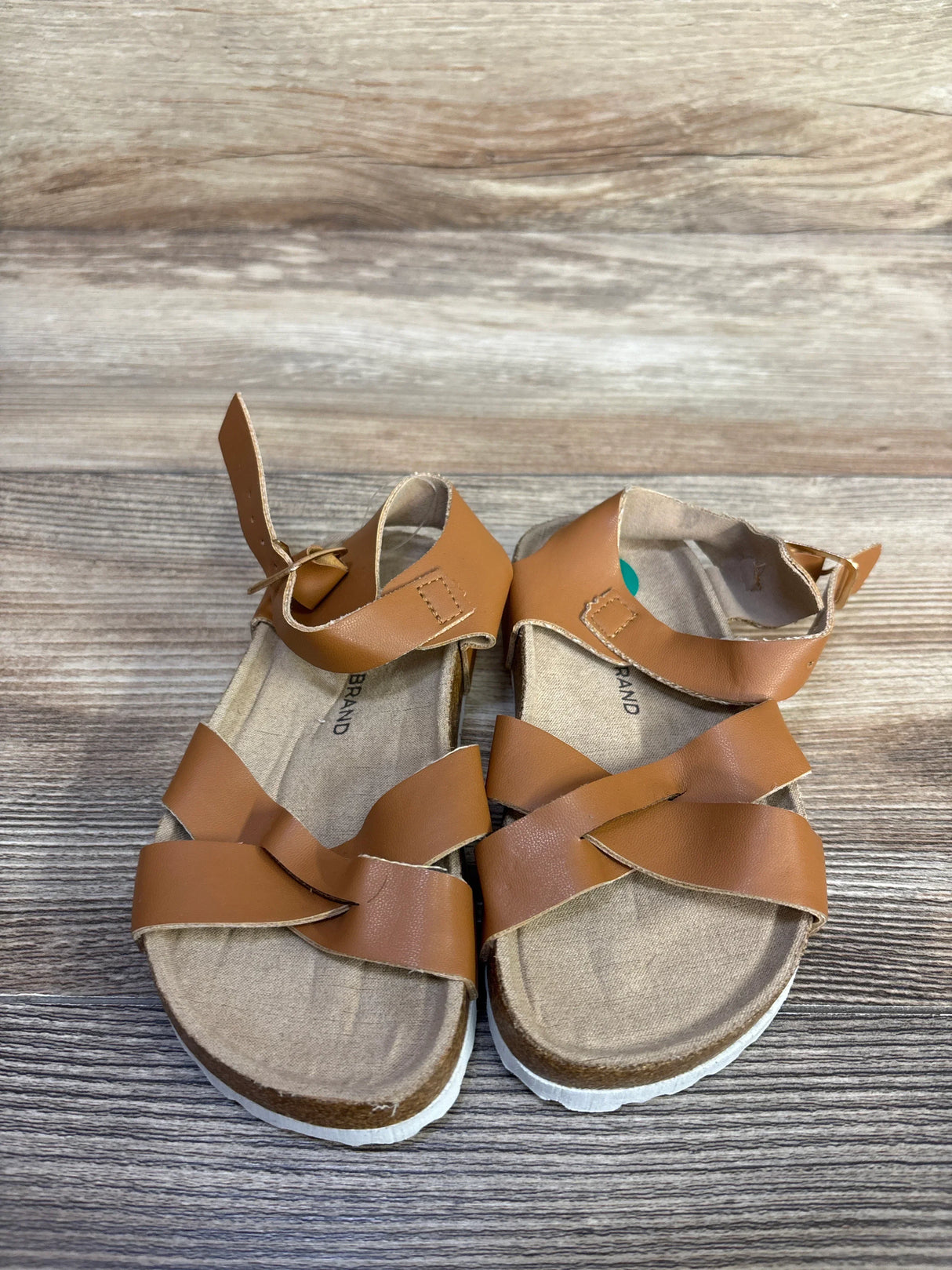 Top-down view of Lucky Brand Kiara toddler sandals, brown; crossed straps, ankle Velcro, contoured footbed on wood background