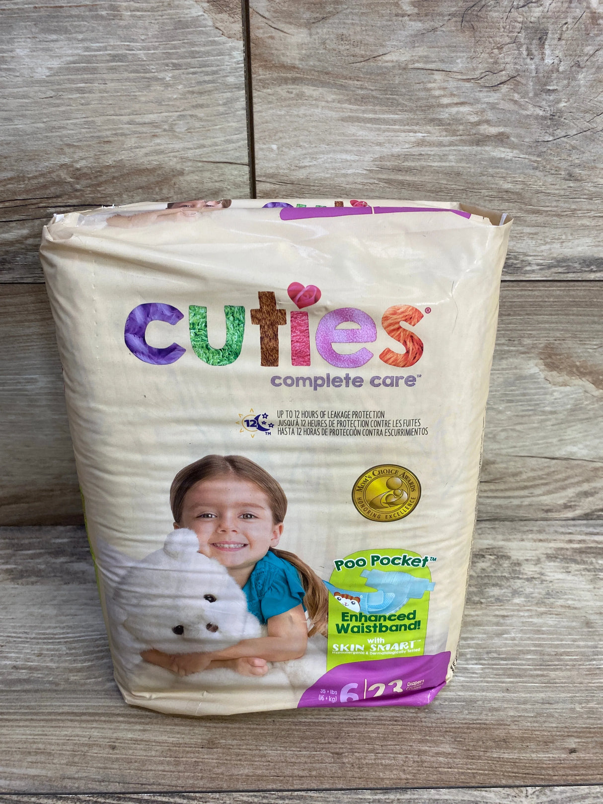 Cuties complete care diapers size 6 pack with child hugging white stuffed animal on wood background