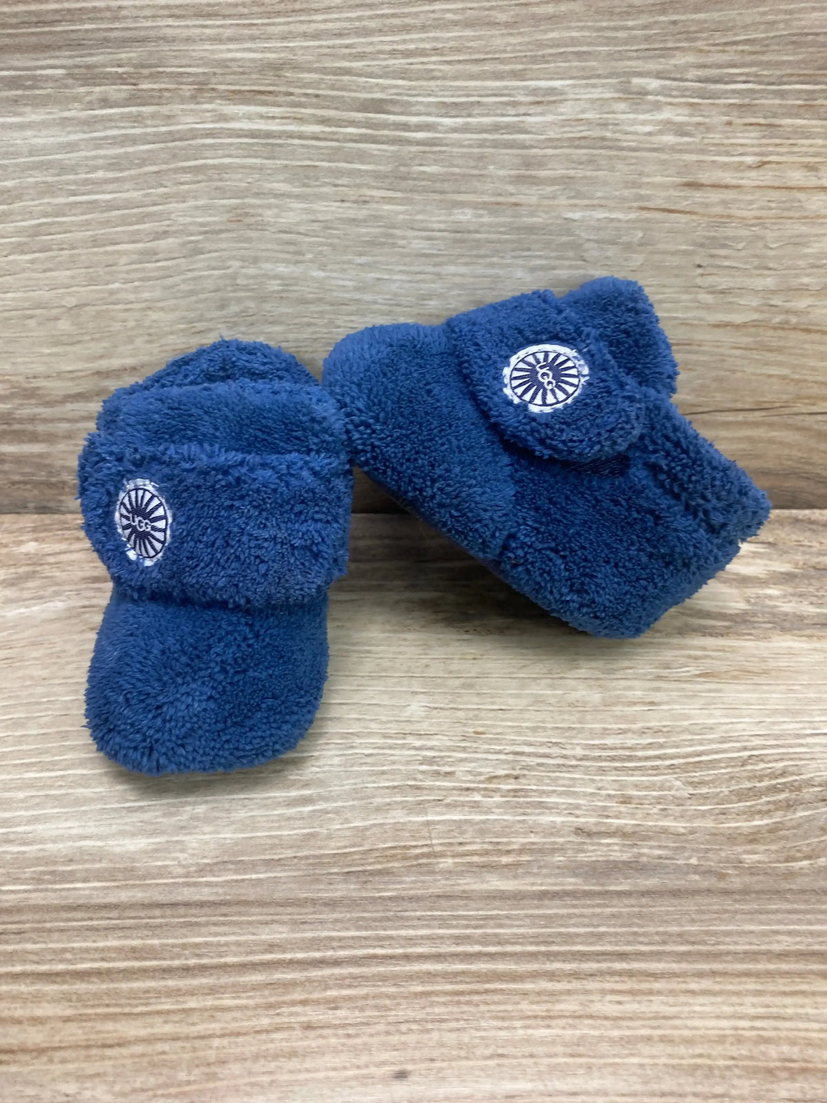 Three-quarter view of UGG Baby Bixbee boots showcasing navy blue plush fleece and fold-over cuff with UGG logo on light wood background