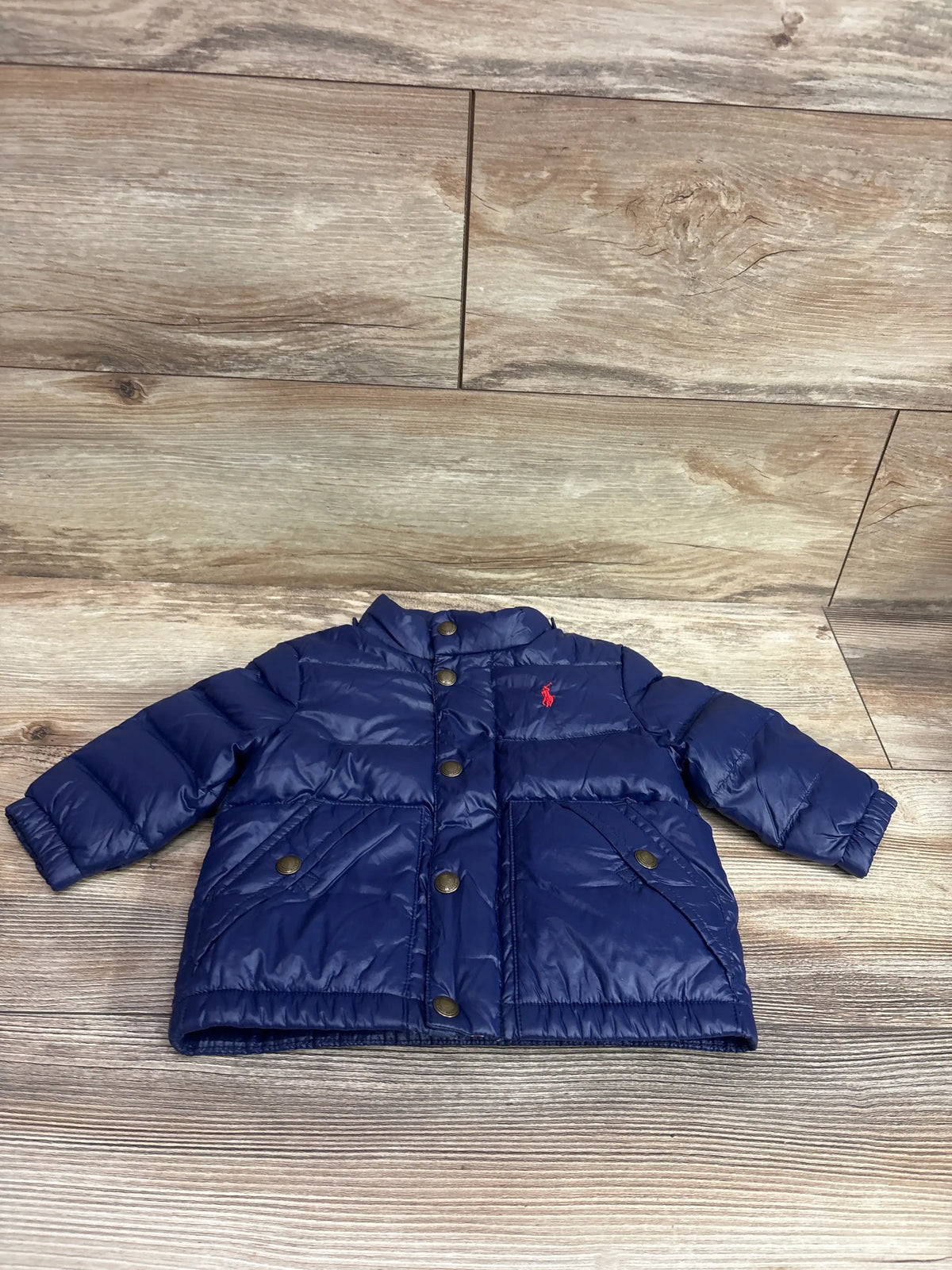 Front view of navy Ralph Lauren puffer jacket, size 9 months, with quilted panels, snap buttons, and pockets on a wood floor