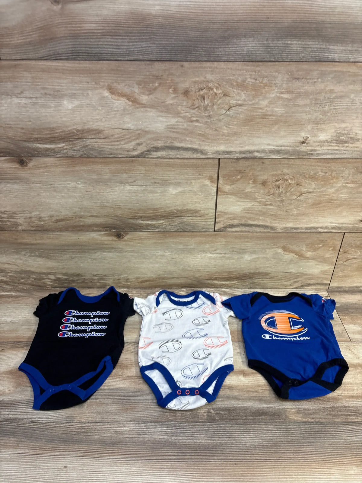 Front view of Champion 3-pack bodysuit set in size 3 months, showing black, white patterned, and blue bodysuits on a wood floor