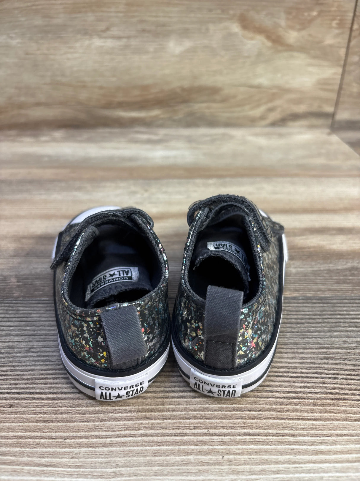 Rear view of size 6C Converse All Star sneakers, glittery black, showing multicolor glitter and heel logo on wood background
