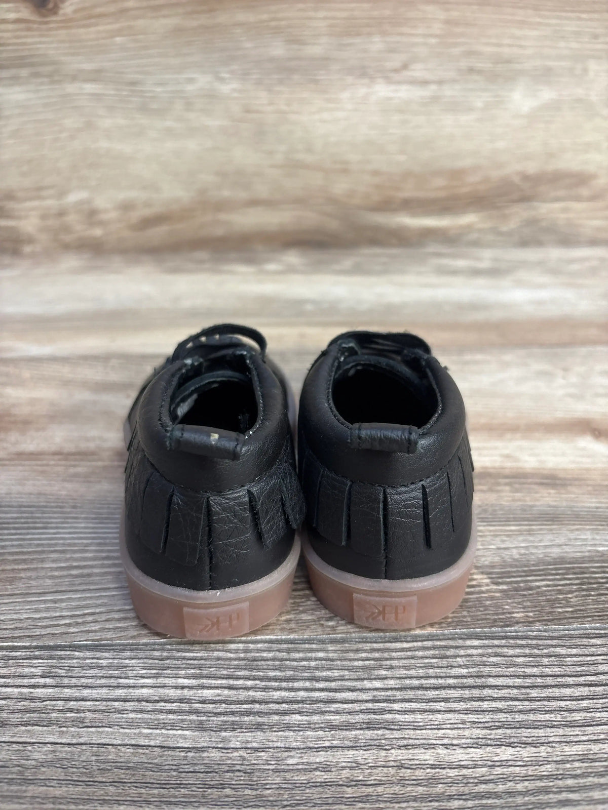 Rear view of Freshly Picked slip-on mid toddler sneakers in black showcasing fringed heel and gum-colored sole on wood plank background