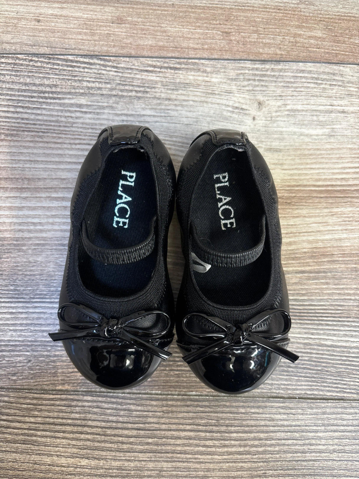 Top-down view of Children's Place black Mary Jane flats with glossy patent toe caps, bow accents and elastic strap on wood floor