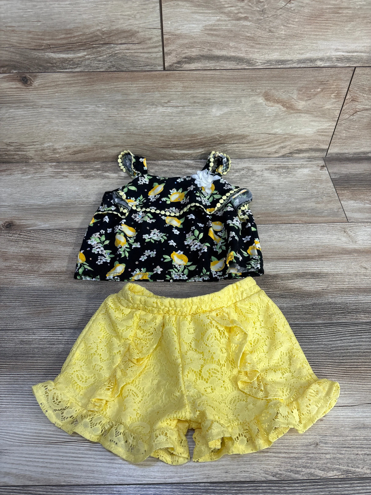 Flat lay of Little Lass 2-piece lemon-print gem top and yellow lace ruffle shorts on wood background