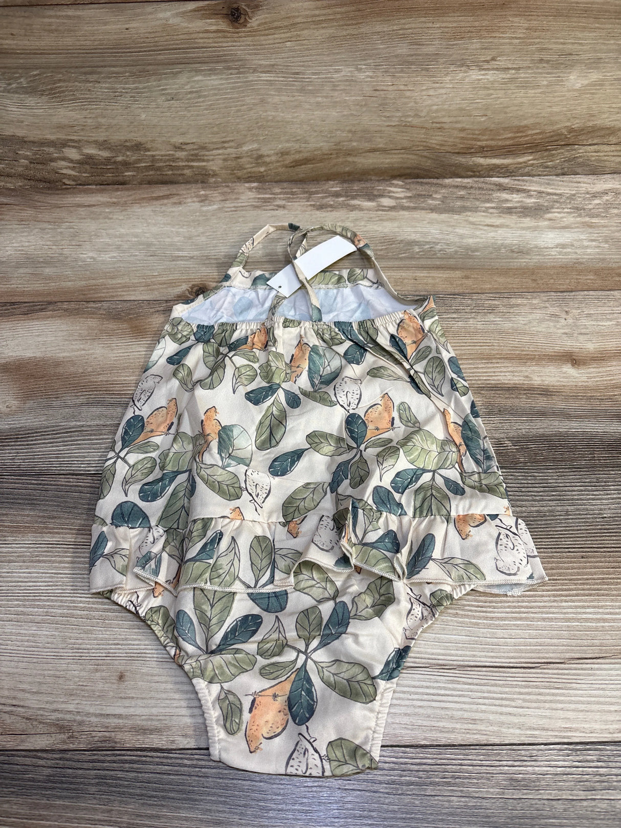 Flat lay of beige leaf-print skirted bubble romper for 18-24 month toddler showing ruffled skirt overlay, adjustable shoulder straps, and elastic leg openings on wood background