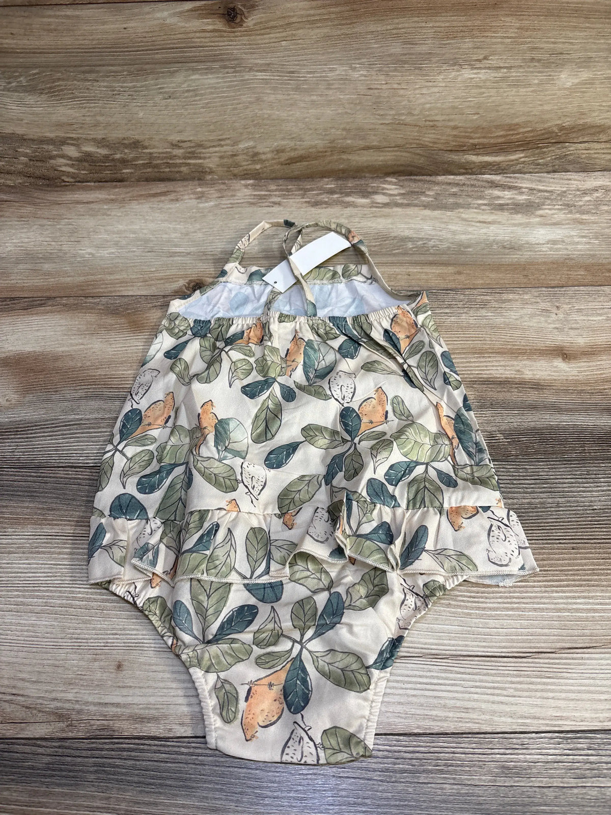 Flat lay of beige leaf-print skirted bubble romper for 18-24 month toddler showing ruffled skirt overlay, adjustable shoulder straps, and elastic leg openings on wood background