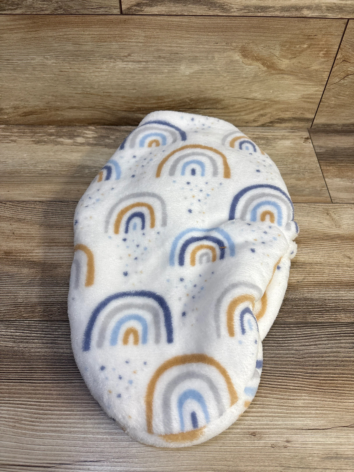 Top-down view of plush swaddle wrap showcasing blue and tan rainbows on white plush fabric with wood background