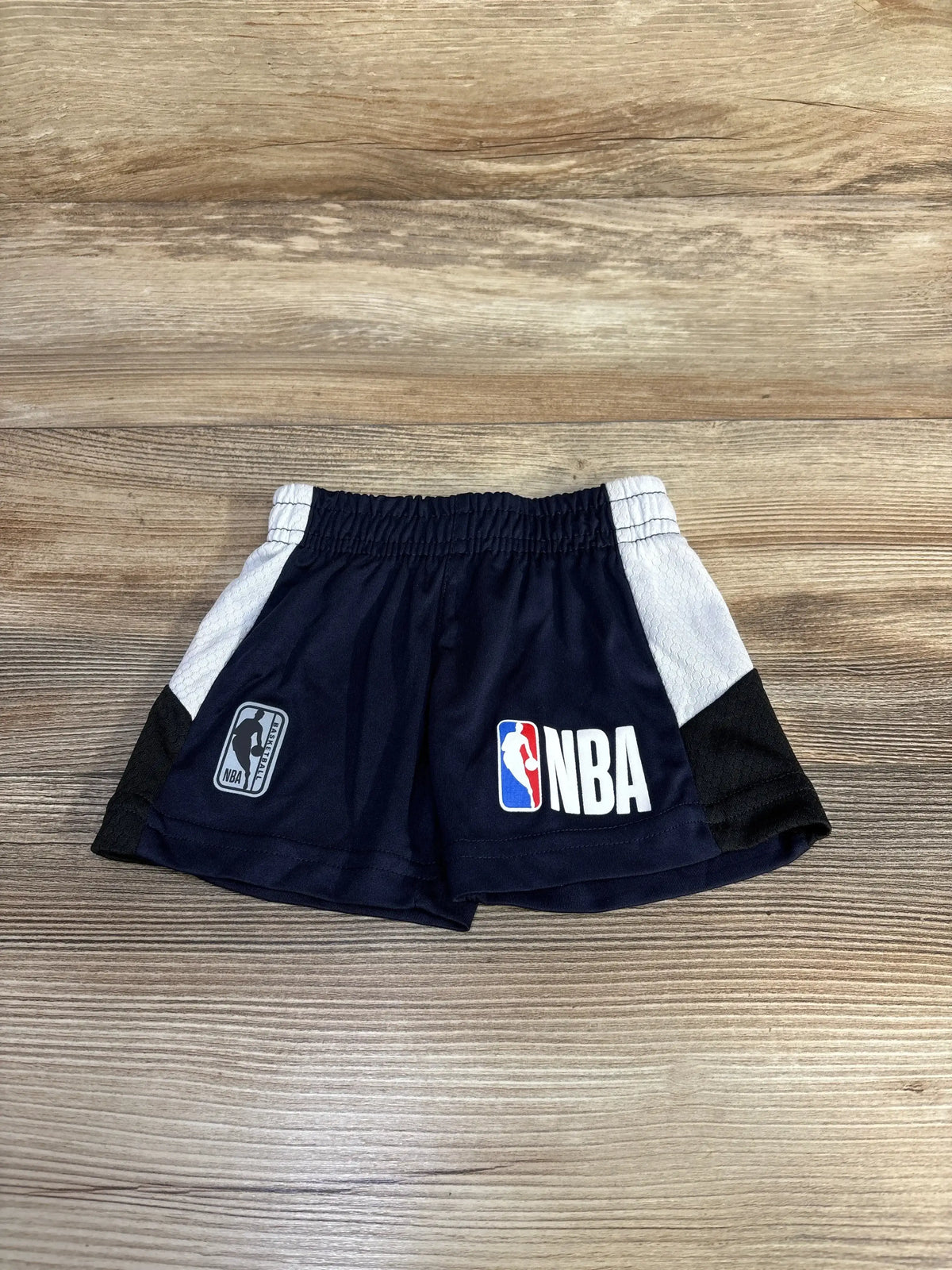 Front flat-lay of navy baby NBA pull-on shorts (3-6 months) with elastic waistband, white side panels and NBA logo on wood