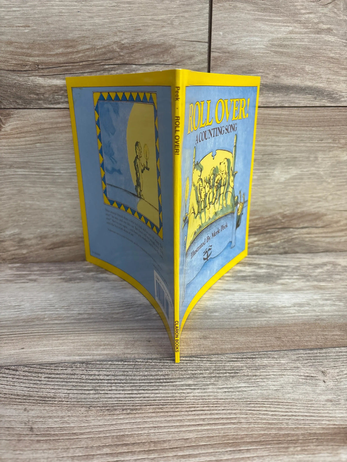 Three-quarter view of Roll Over! paperback showing illustrated blue-and-yellow cover with stacked animals and title ‘ROLL OVER! A COUNTING SONG’ on a wood plank background