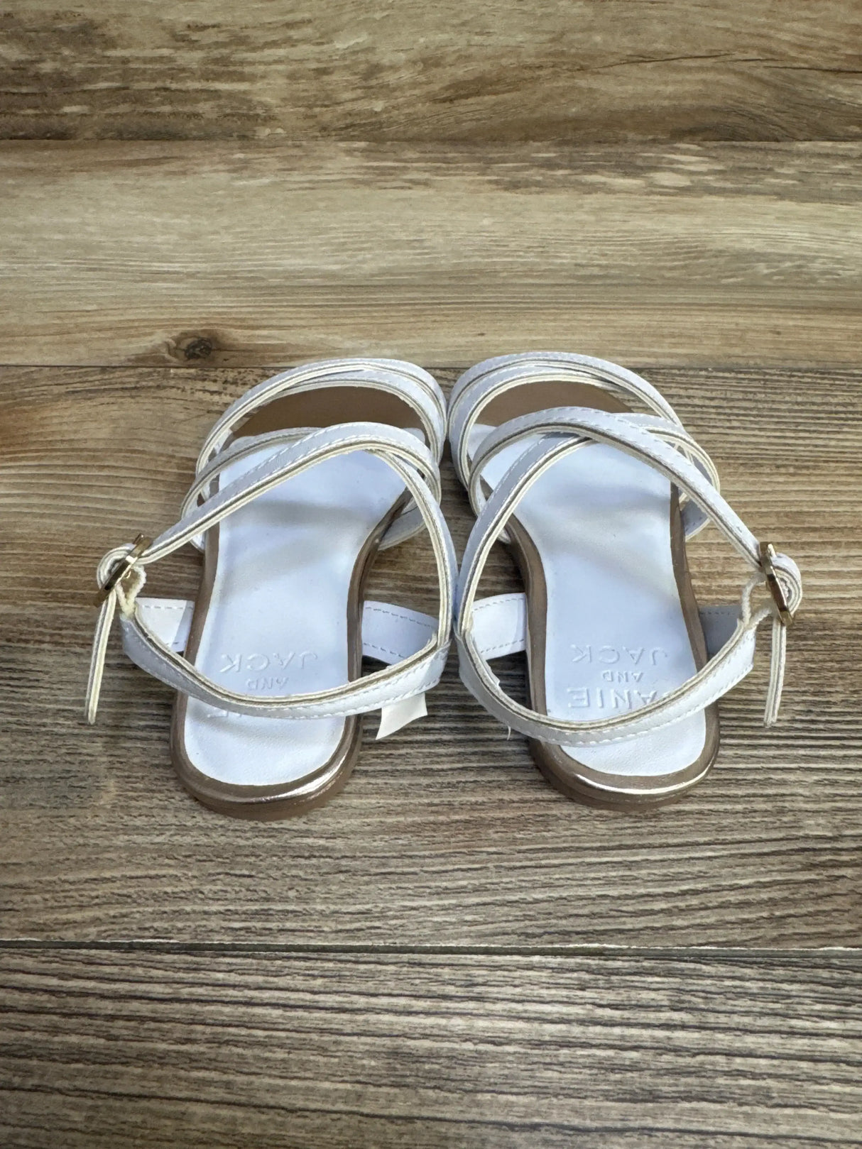 Top view of Janie & Jack white metallic trim strap sandal size 7C with crossed straps and ankle buckle on wood floor