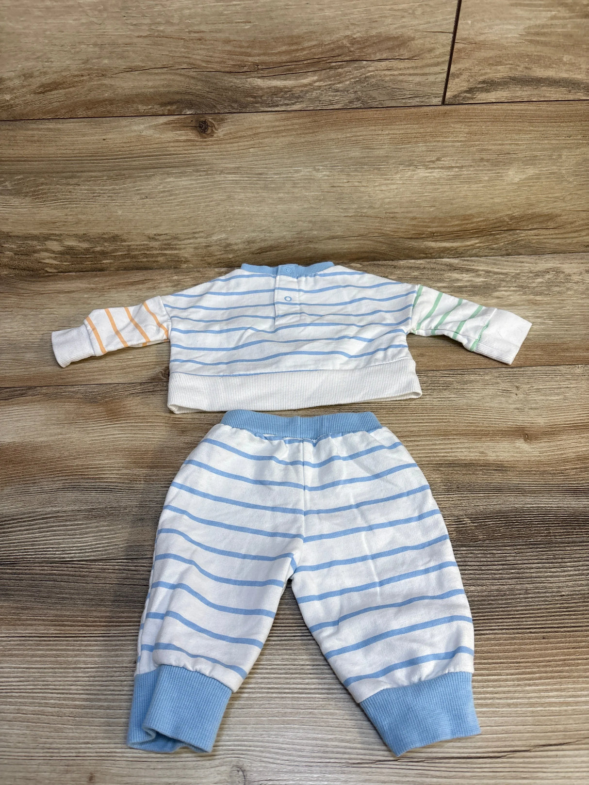 Flat-lay of Disney Baby Mickey Mouse newborn 2-piece long-sleeve shirt and joggers showing light-blue horizontal stripes, mismatched orange and green sleeves, and blue ribbed cuffs on wood background