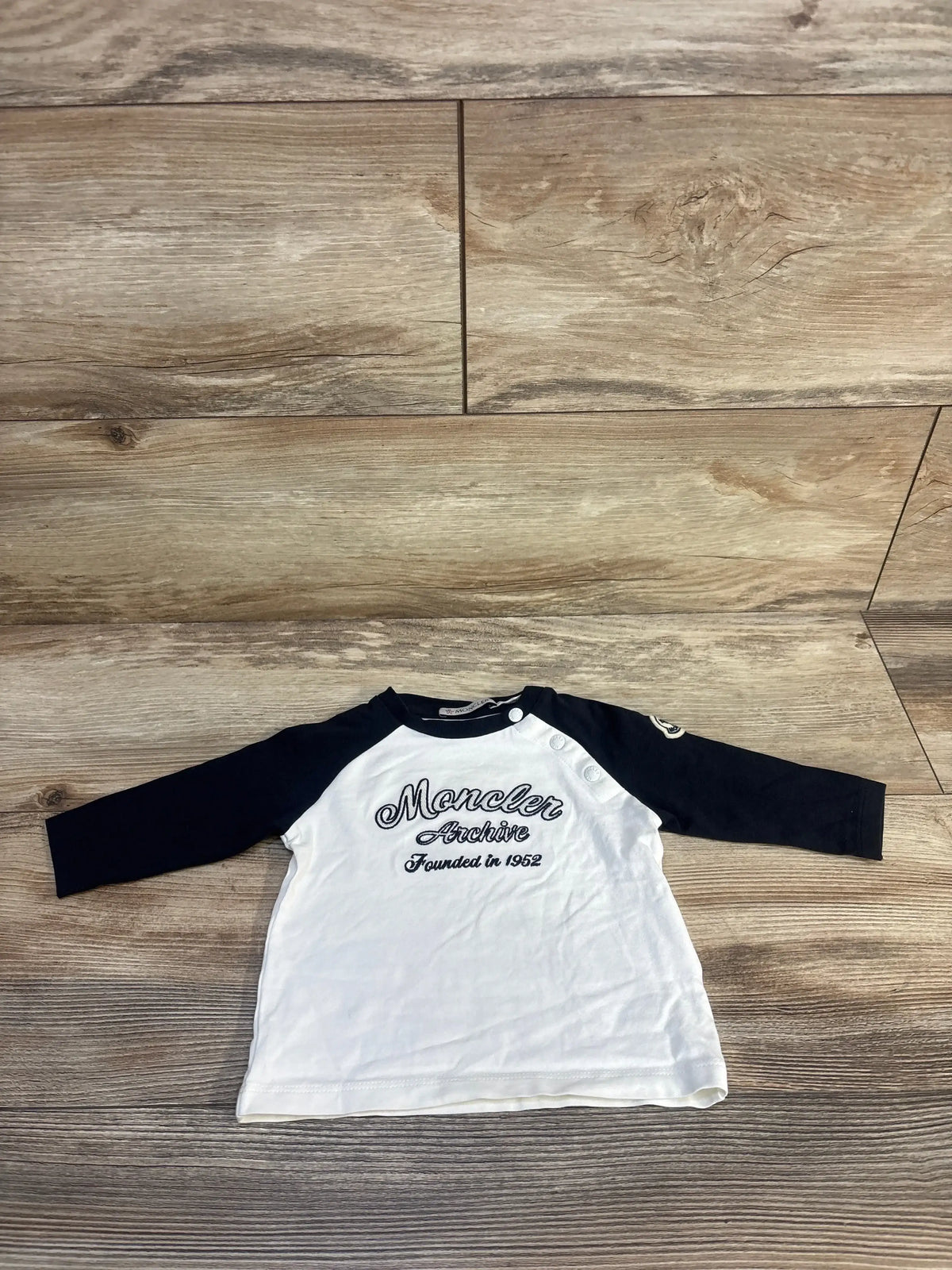 Flat lay of Moncler raglan shirt with navy sleeves, white body, chest text Moncler Archive 1952, shoulder snaps on wood floor