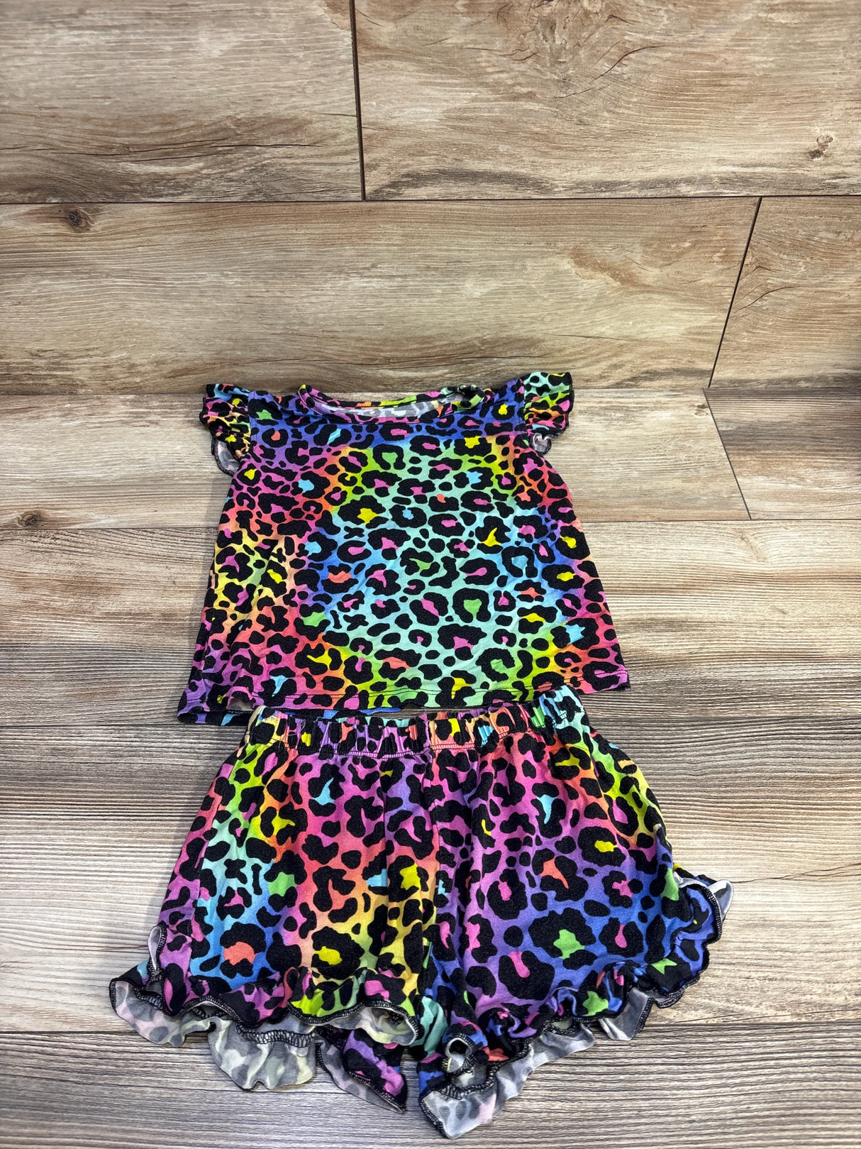Top view of Little Snugglers bamboo 2-piece toddler pajama set, rainbow leopard print with ruffle sleeves and ruffled shorts on wood