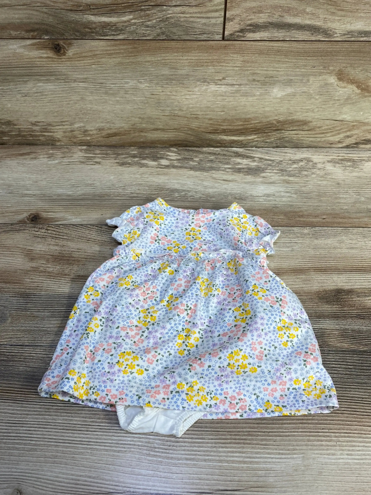 Flat lay of Just One You 2-piece white baby floral dress and matching bloomers showcasing ruffle sleeves and gathered skirt on wood background