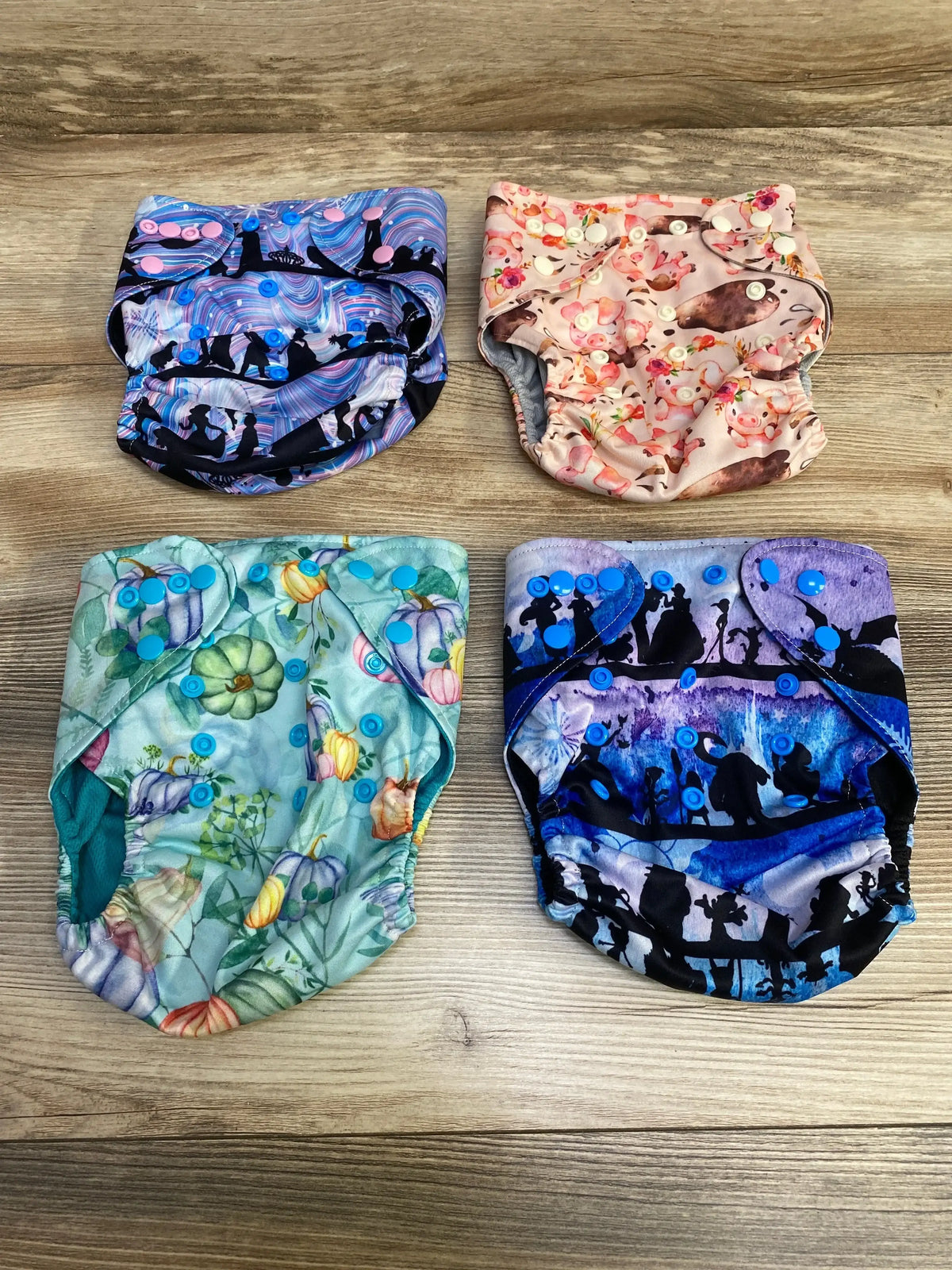 Flat lay of Happy BeeHinds 4 Pack Pocket Diapers showcasing four printed cloth diapers in pink floral teal and purple fairytale prints on wood plank floor
