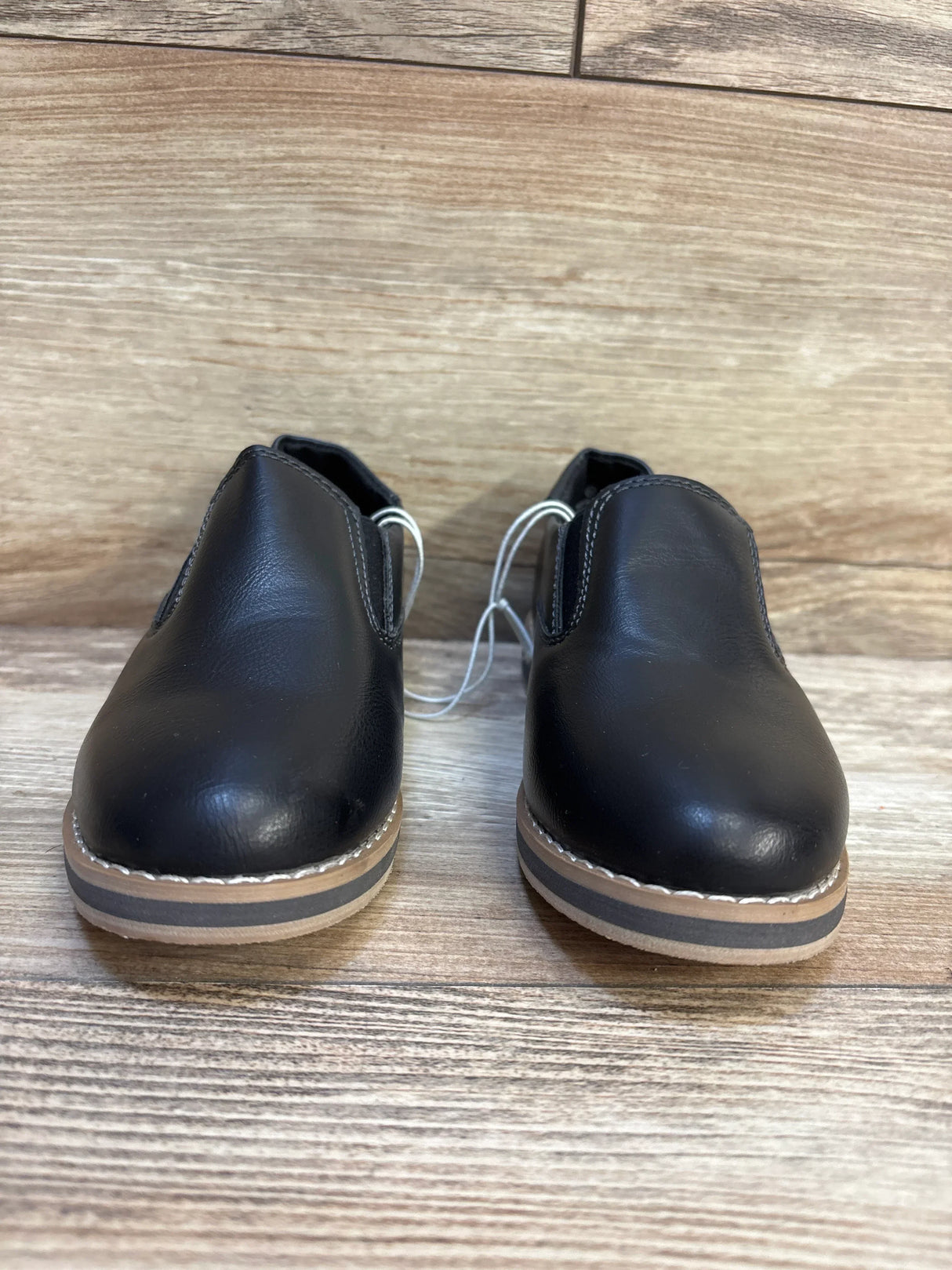 Front view of pair of black slip-on kids dress shoes showcasing smooth leather upper striped tan and grey soles on wood plank floor