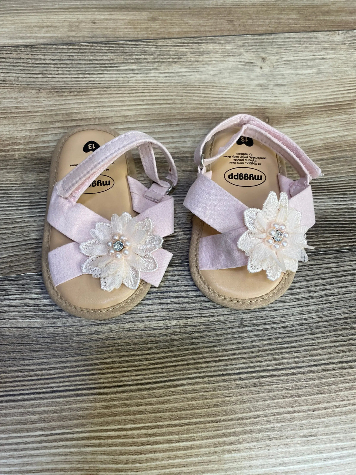Top view of pink baby cross-strap sandals showcasing pearl flower embellishments and Velcro ankle straps on wood floor