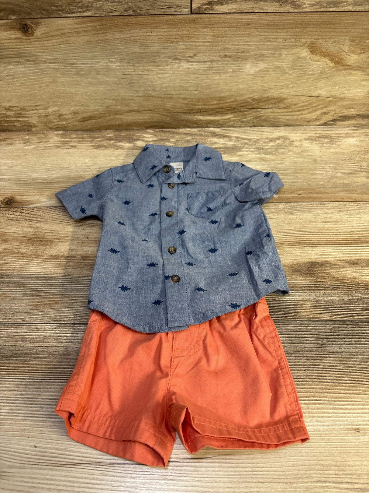 Flat lay of two-piece baby button-up set with blue printed shirt and orange elastic-waist shorts on wood background