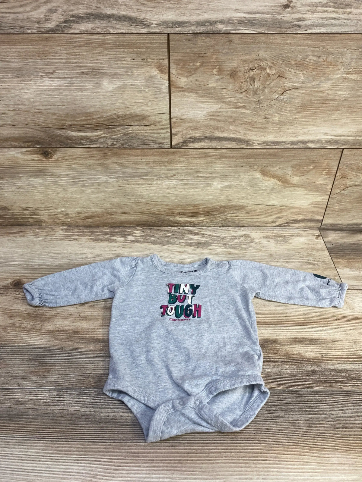 Flat lay of grey Carhartt long-sleeve baby bodysuit (6 months) showcasing multicolor TINY BUT TOUGH chest print on wood floor