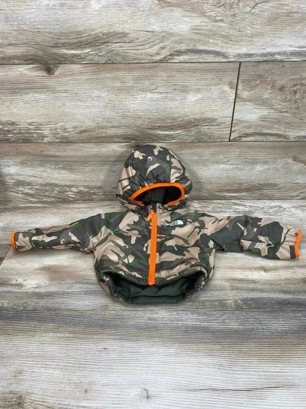 Front view of baby The North Face reversible Perrito jacket showing green camo print, orange zipper, hood on wood background