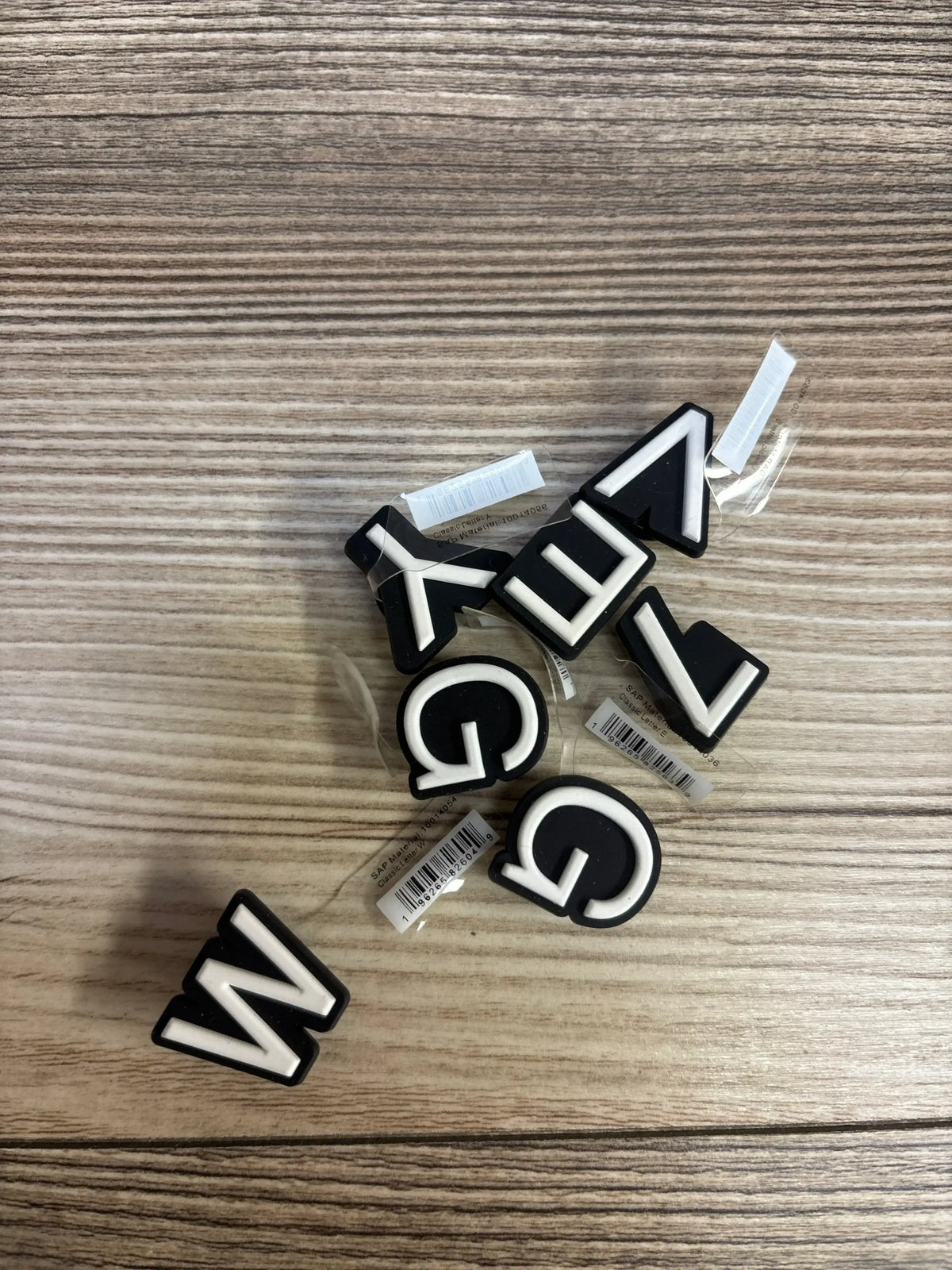 Overhead view of Crocs Jibbitz letter charms showcasing black and white alphabet letters and barcode price tags on a wood grain surface