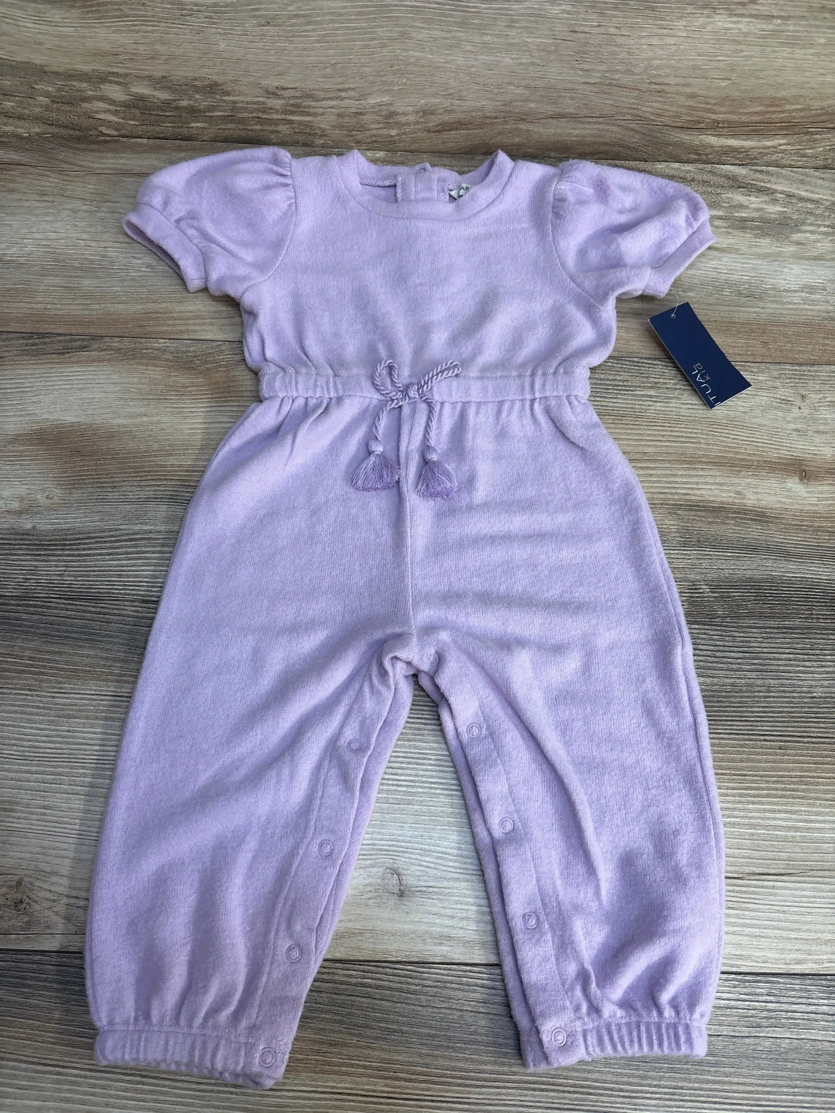 Flat lay of purple short-sleeve baby jumpsuit showing drawstring waist, snap leg closures, and elastic cuffs on wood background