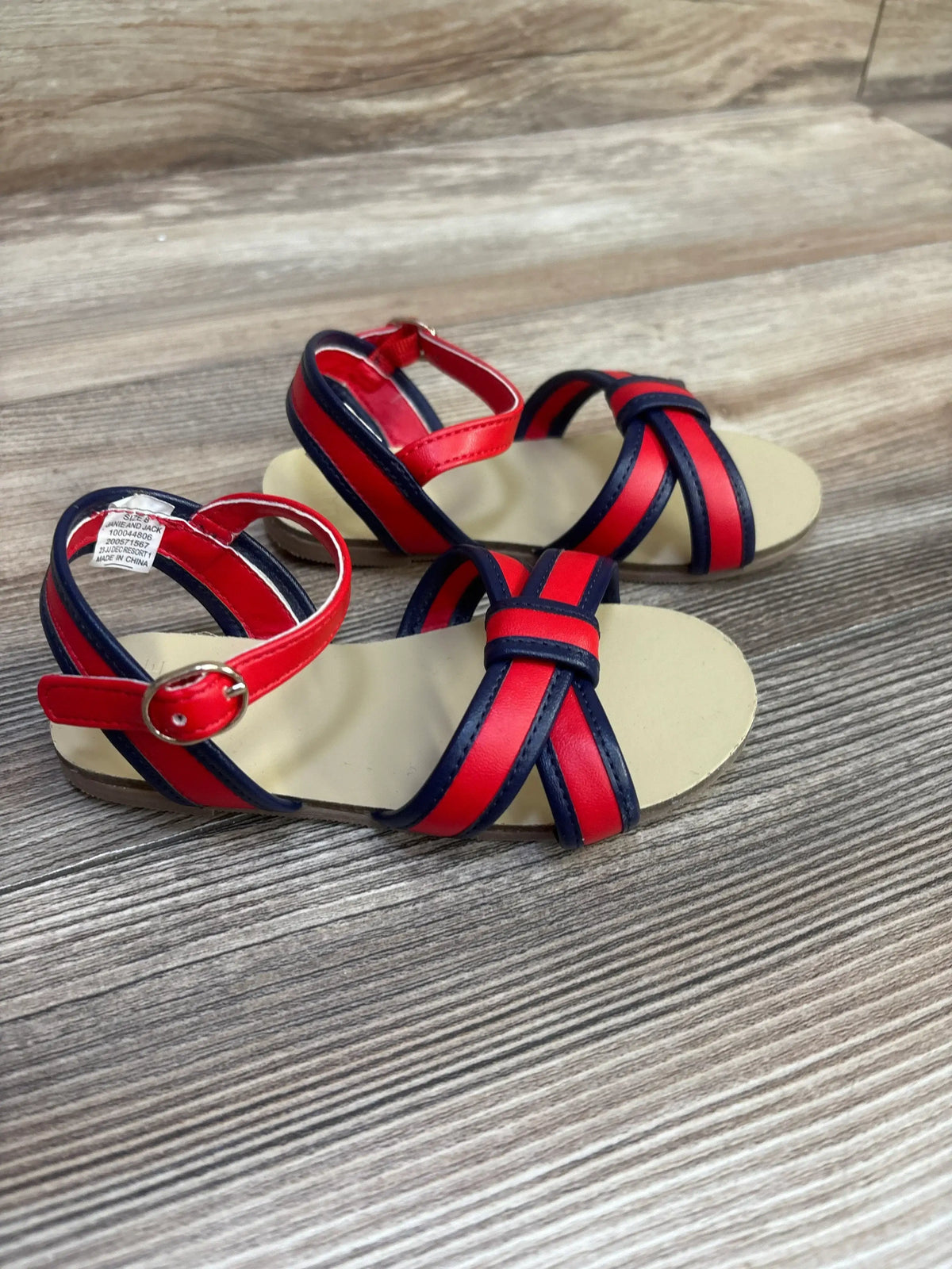 Three-quarter view of Janie & Jack toddler sandals with red and navy crossed straps and ankle buckle on wood floor