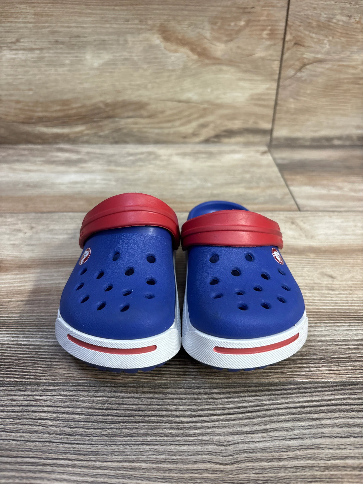Front view of Crocs Crocband II toddler clogs, blue with red heel straps and white soles with red stripe on wood backdrop