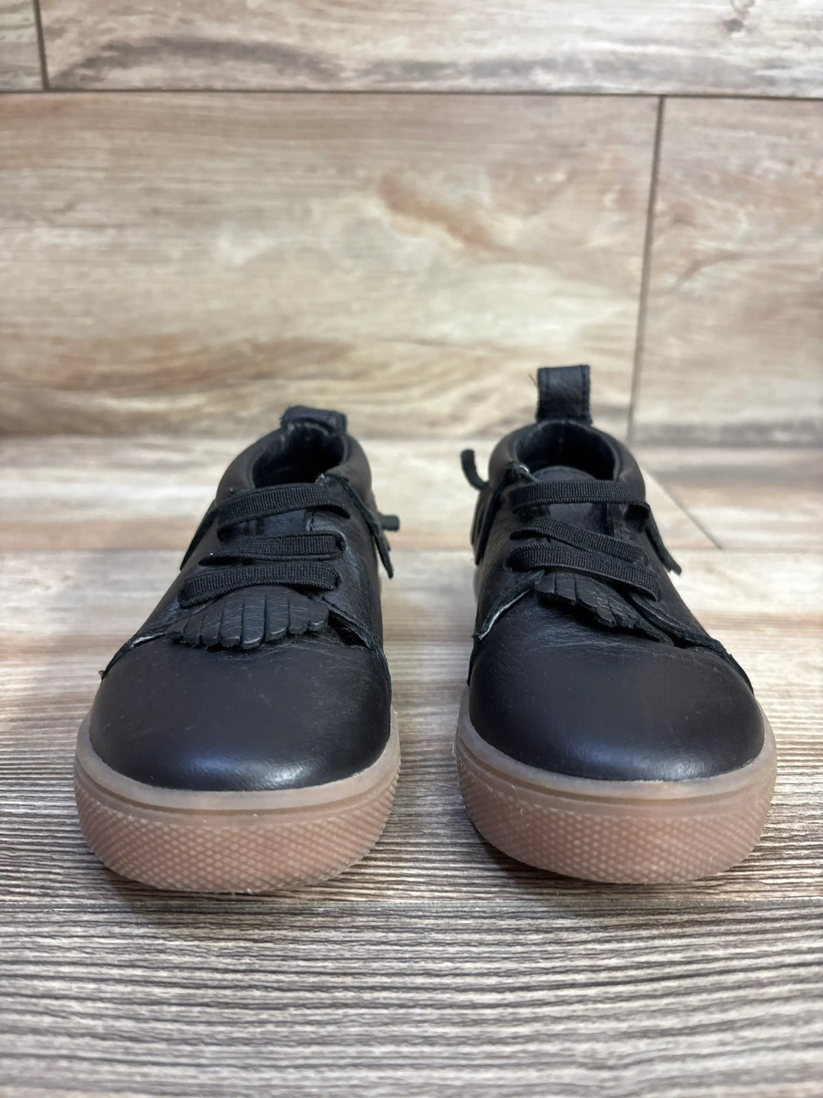 Front view of Freshly Picked Ebony Mocc sneakers showing black leather, fringe kiltie and brown rubber soles on wood floor