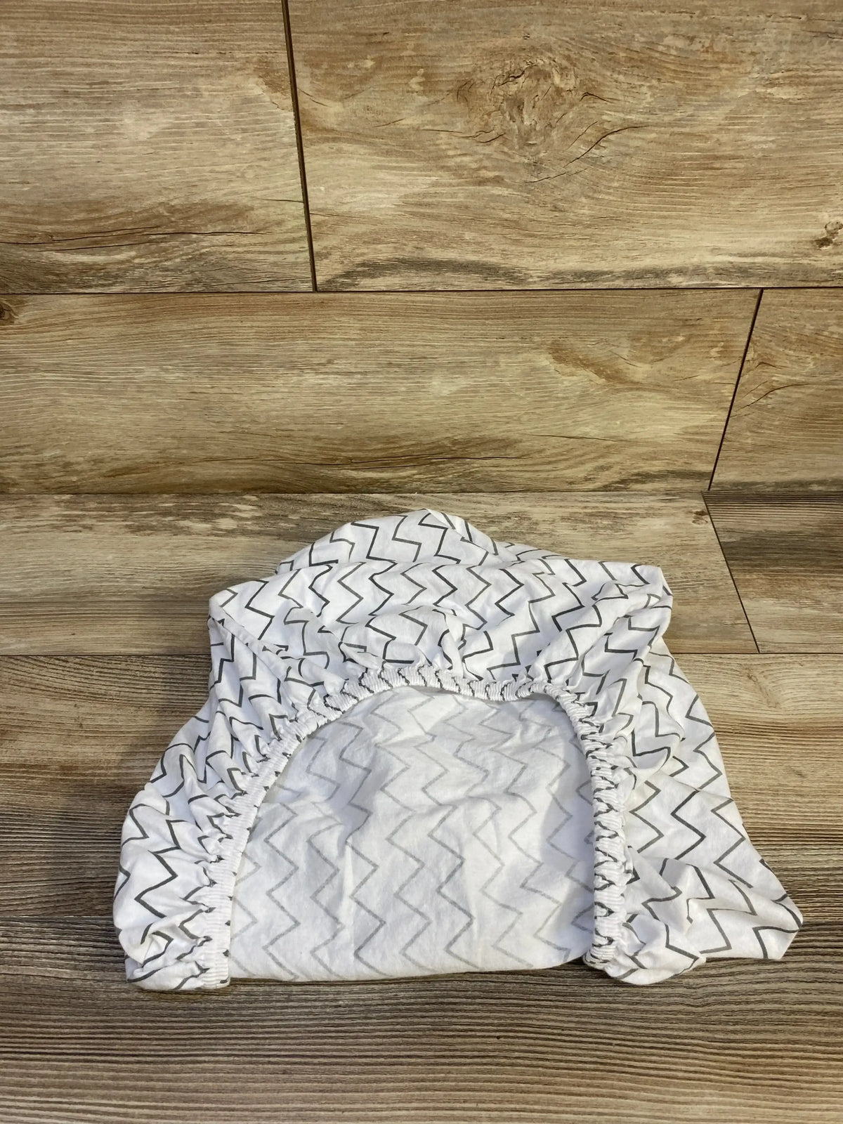 Angled top view of TL Baby fitted bassinet sheet showing grey chevron print and elasticized fitted edge on wood floor background