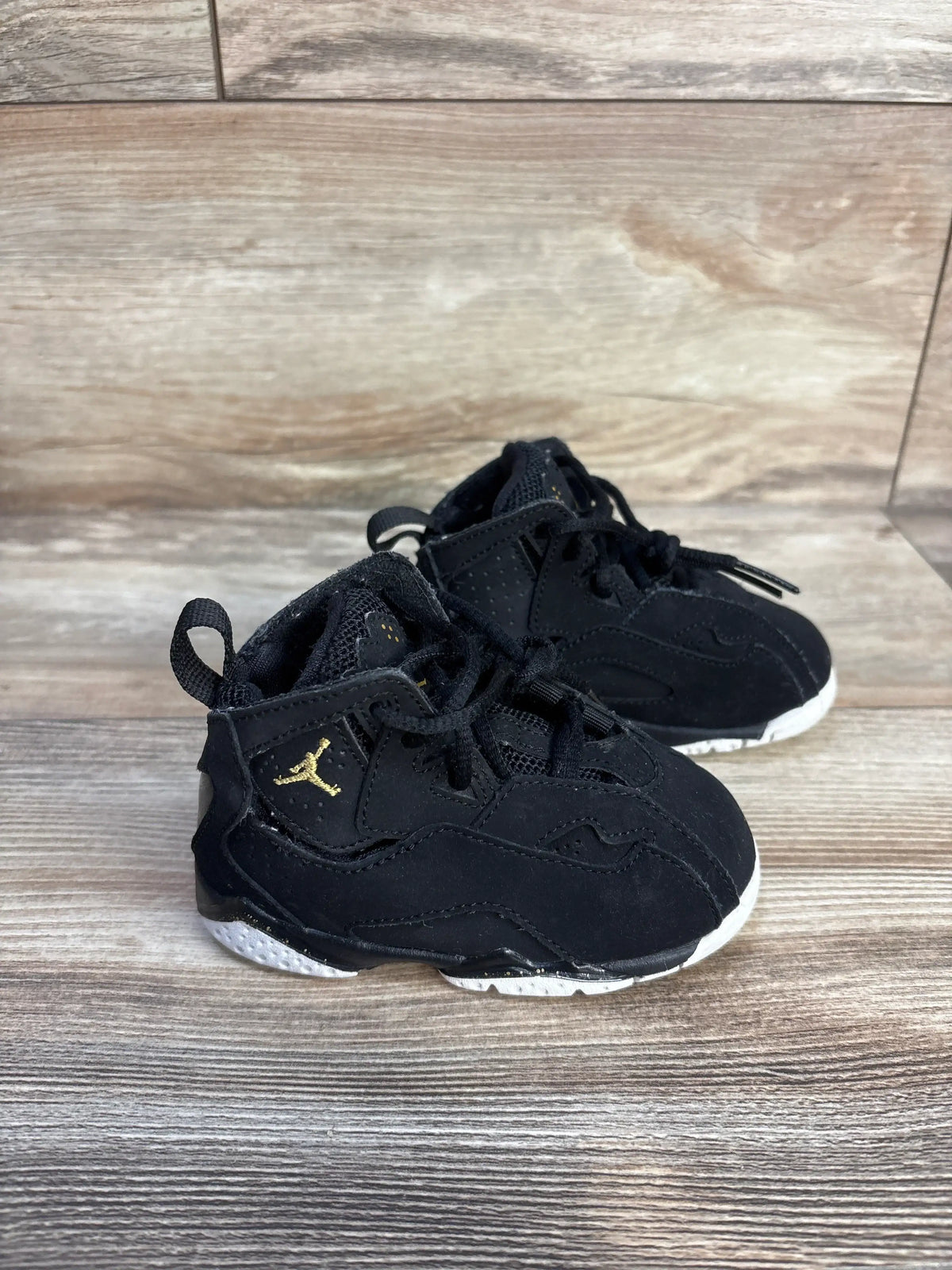 Three-quarter view of black Air Jordan True Flight BT sneakers with gold Jumpman logos and white soles on a wood floor
