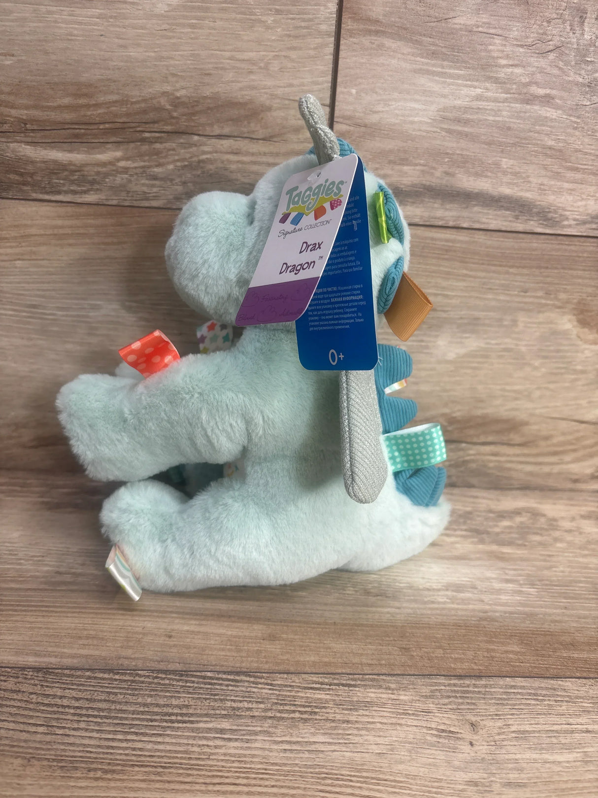 Side view of Taggies Drax Dragon plush in light blue showcasing silver wings ribbon tags and Signature Collection hangtag on wood plank floor