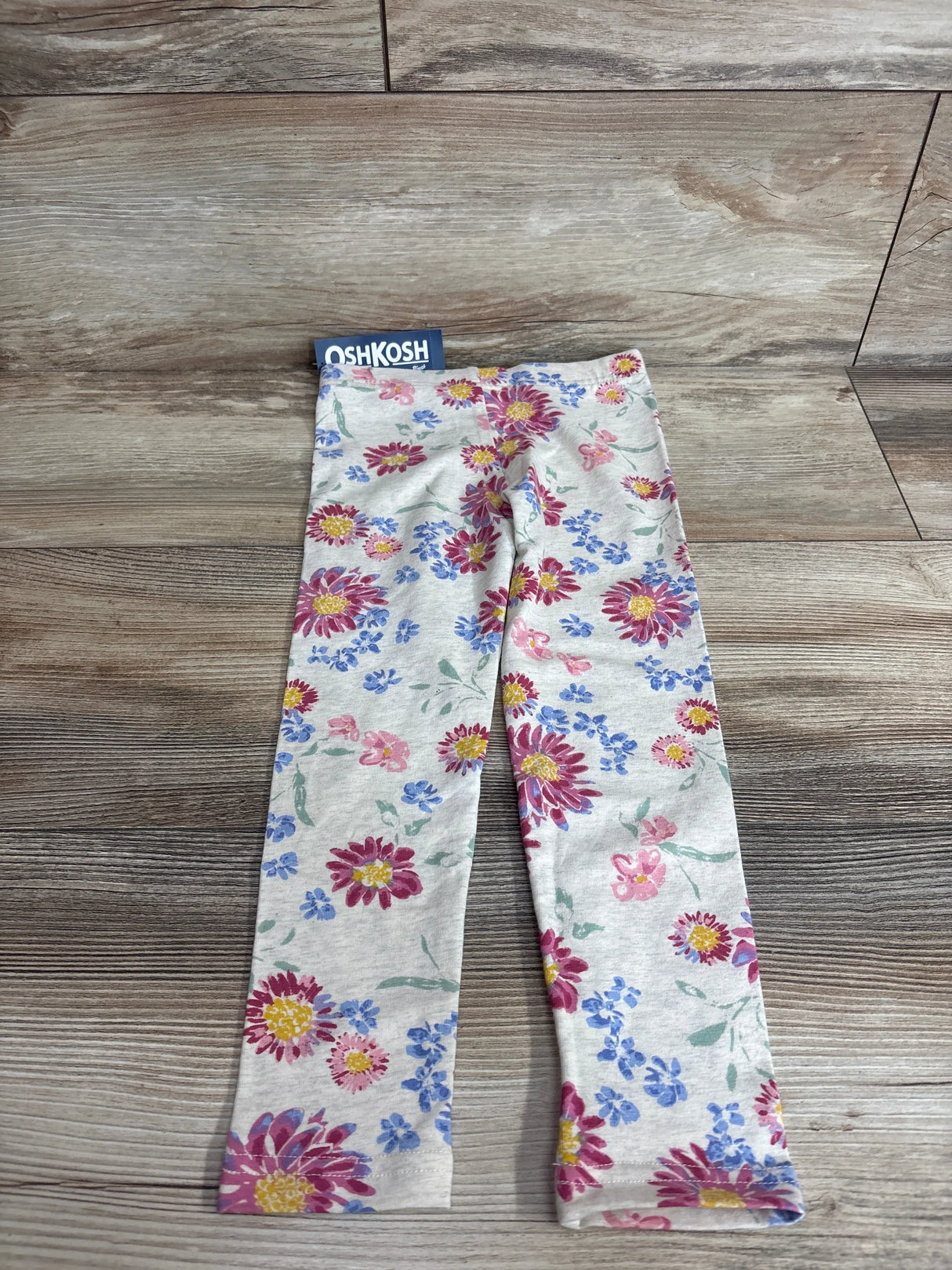 Flat lay of Oshkosh girls’ beige floral leggings size 4T showing full-length pink and blue flower print on wood floor